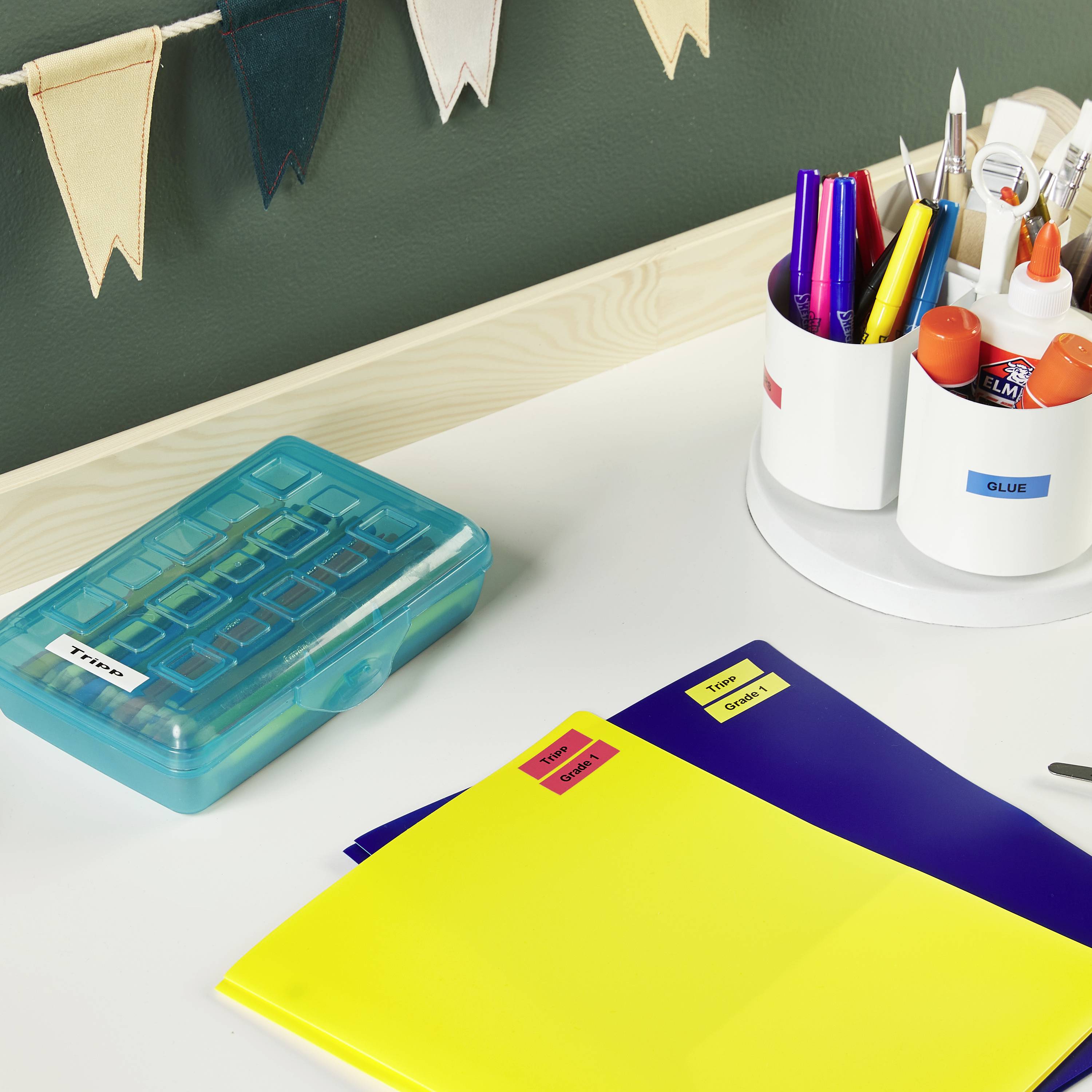 Artykuły biurowe na biurku: niebieskie i żółte segregatory, kolorowe pisaki w pojemniku, pudełko z etykietami oraz proporczyk na ścianie.