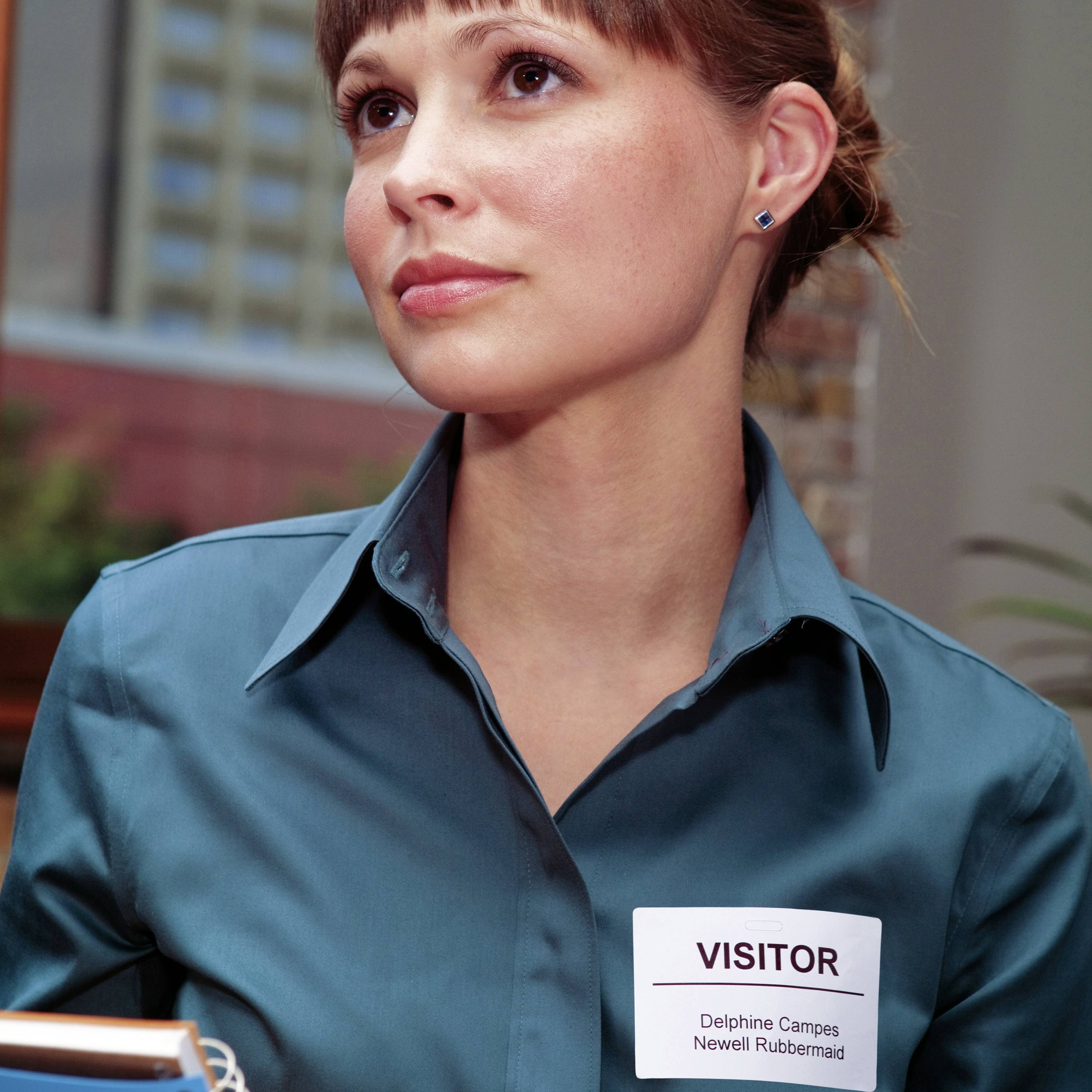 Osoba w niebieskiej koszuli nosi identyfikator z napisem 'Visitor'. Patrzy zamyślonym wzrokiem w bok.