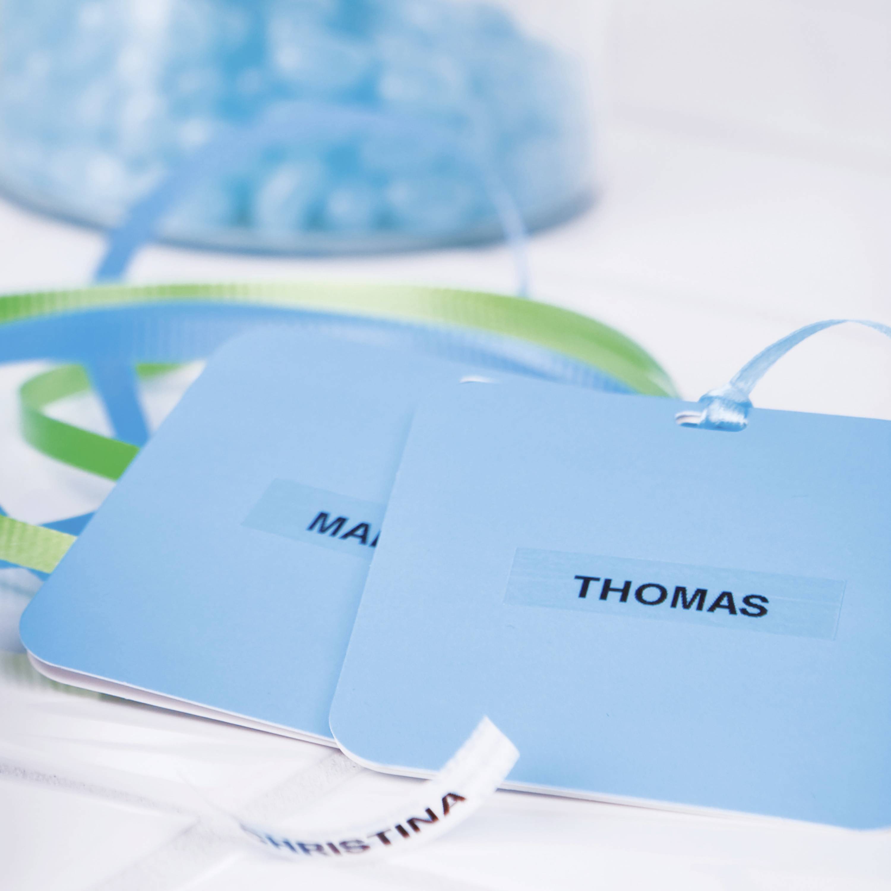 Dwa niebieskie identyfikatory z imionami 'MARIA' i 'THOMAS' leżą na stole. W tle widoczne są niebieskie kamienie szklane.
