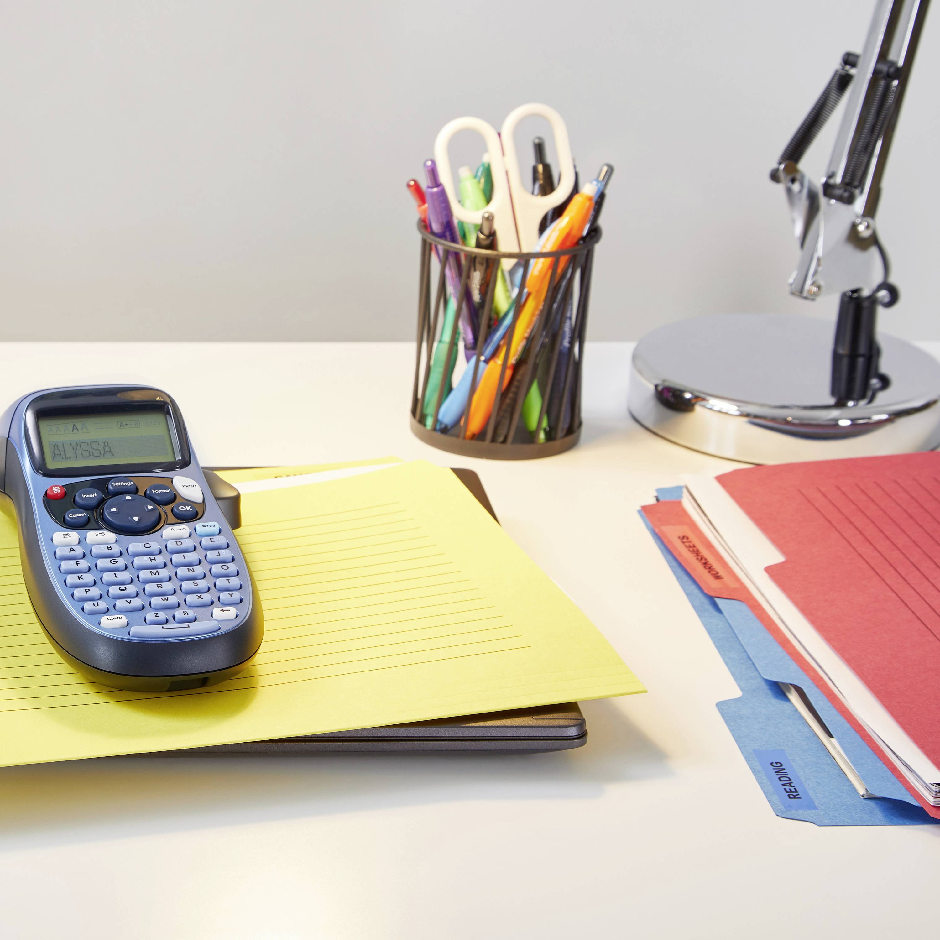 Biurko z drukarką etykiet na żółtej teczce, obok pojemnik na długopisy z kolorowymi piórami oraz kilka segregatorów.