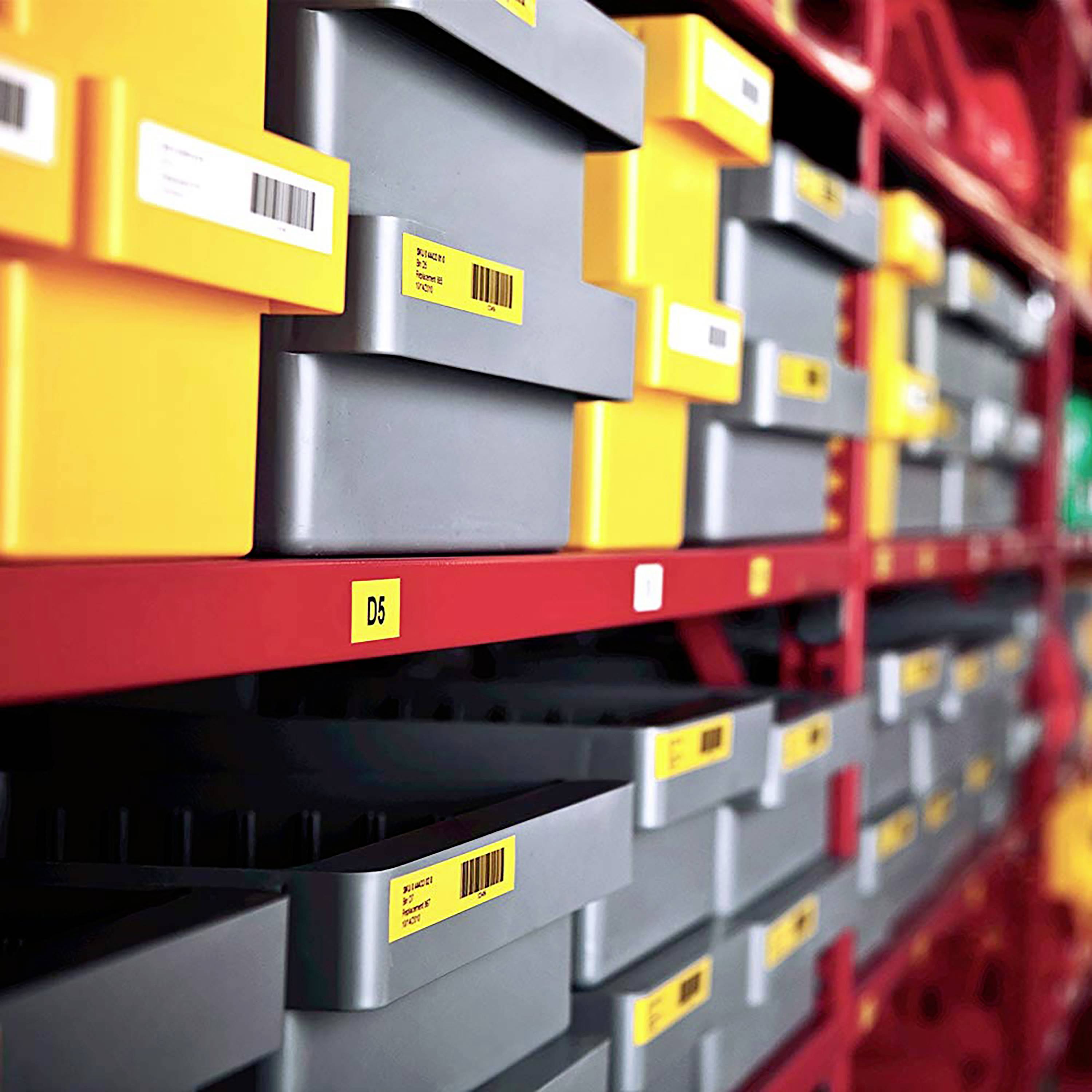Kilka plastikowych pudełek w czerwonym regale, ponumerowanych i oznaczonych kolorowymi etykietami, w magazynie.