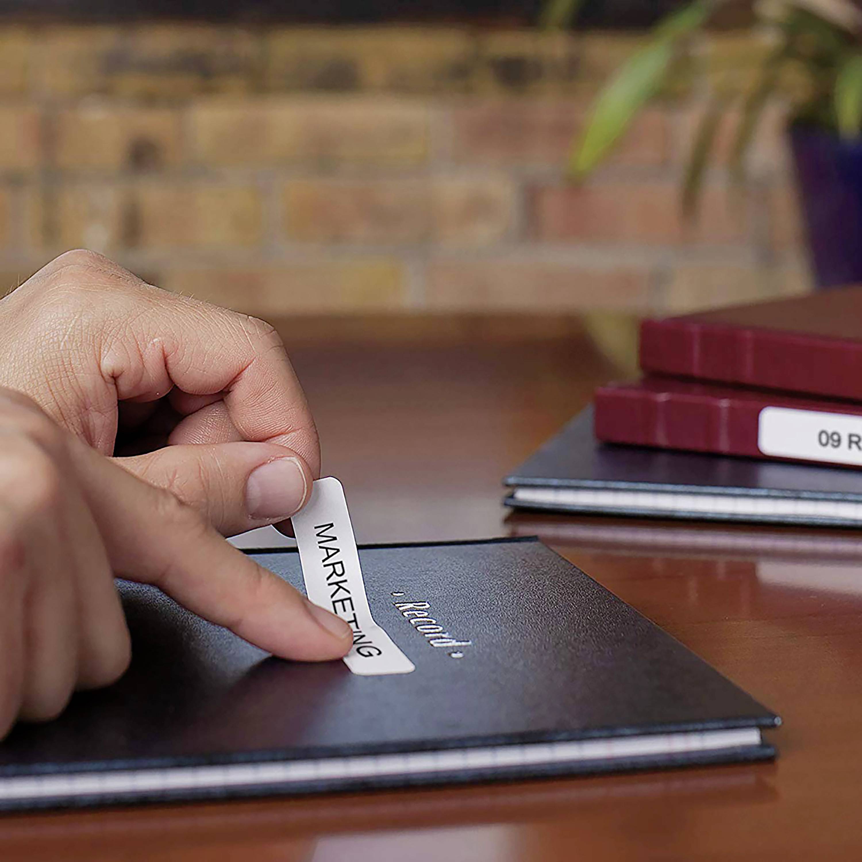 Ręka nakłada naklejkę z napisem 'Marketing' na czarny notes. W tle widoczne są książki i roślina.