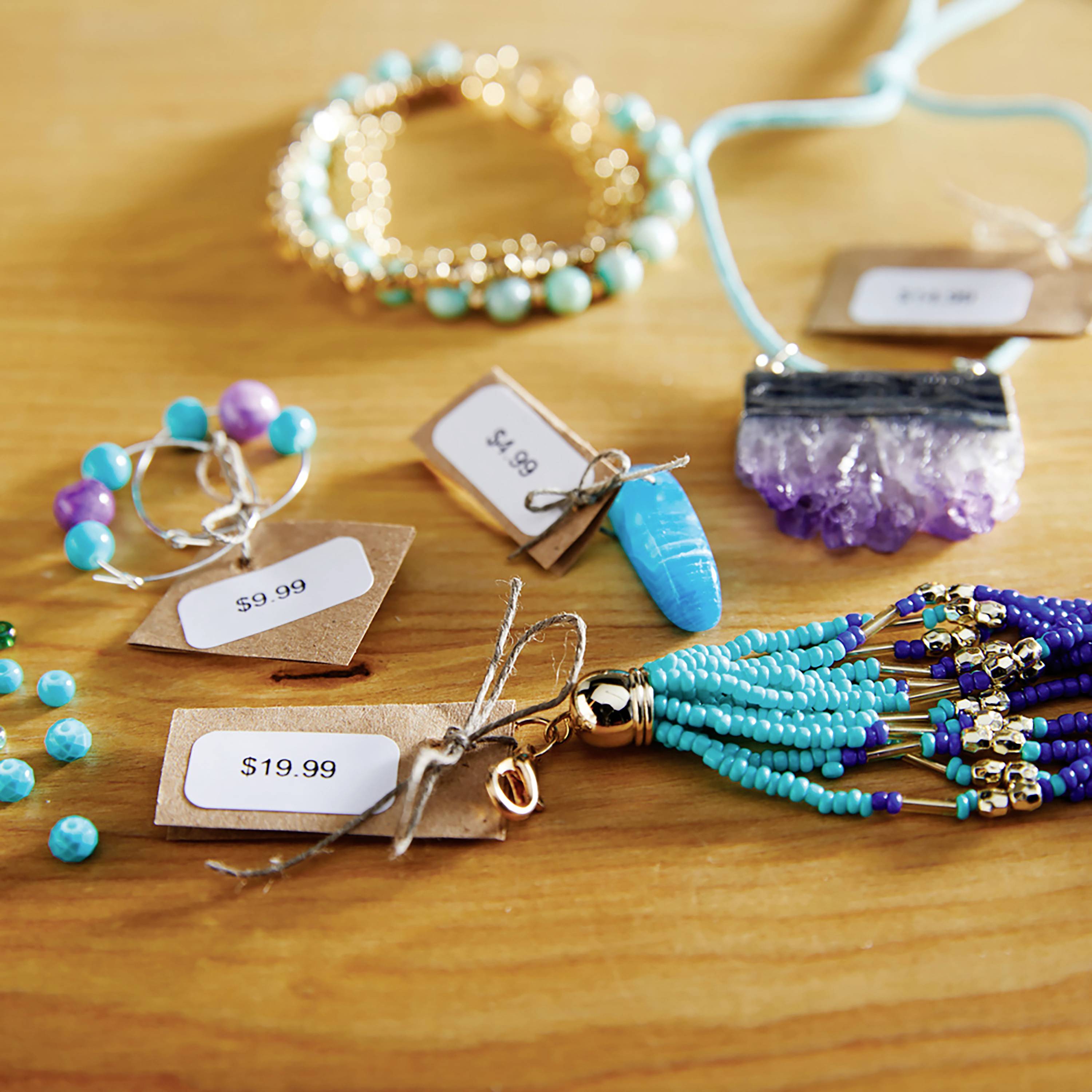 Biżuteria na drewnianym stole: turkusowe i fioletowe koraliki, bransoletki, kolczyki z metkami cenowymi '19,99 <!--ssr-html-->#39;, '8,95 <!--ssr-html-->#39;, '9,99 <!--ssr-html-->#39;.