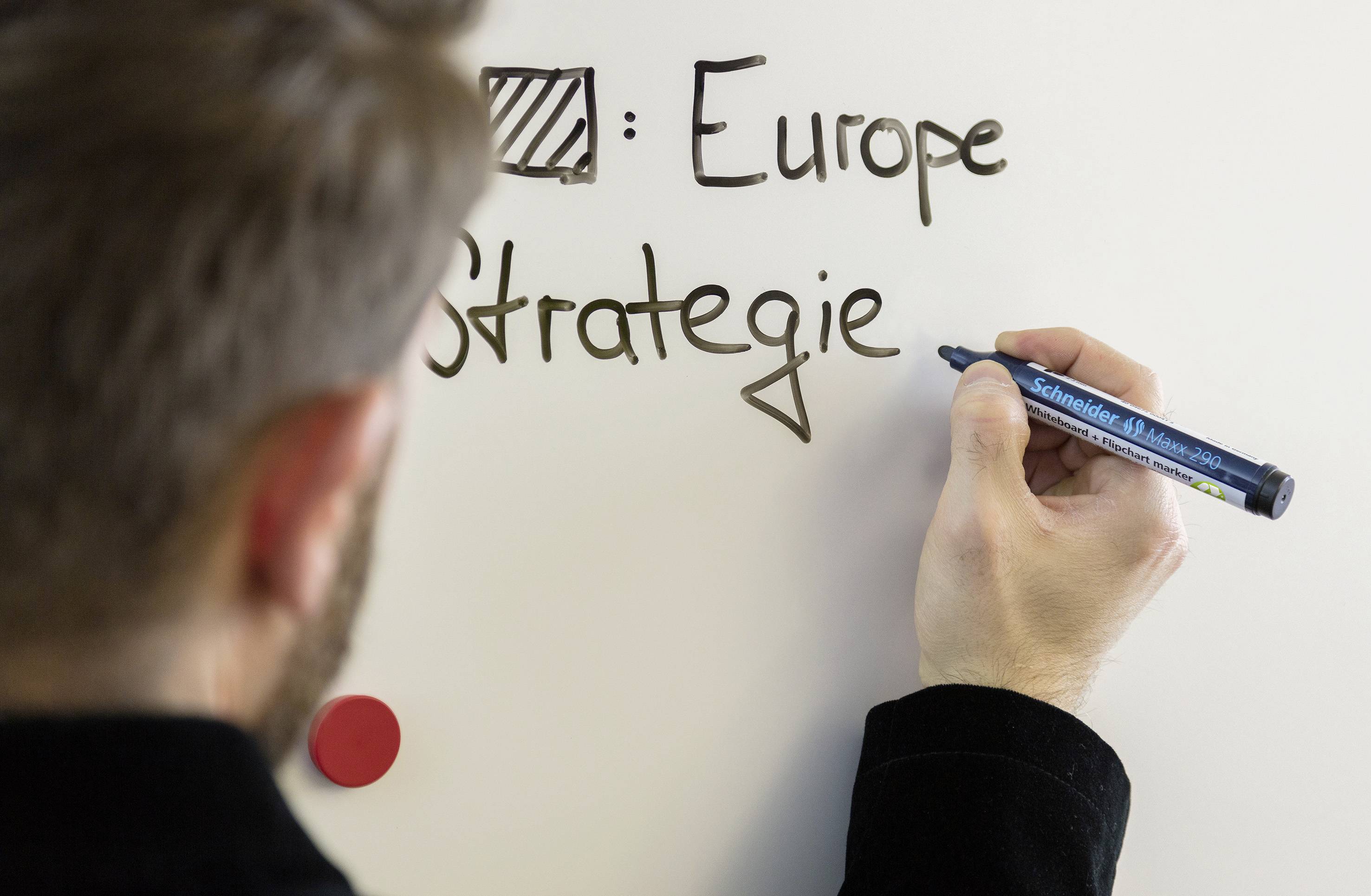 Osoba pisze czarnym piórem słowa 'Europe Strategie' na białej tablicy.