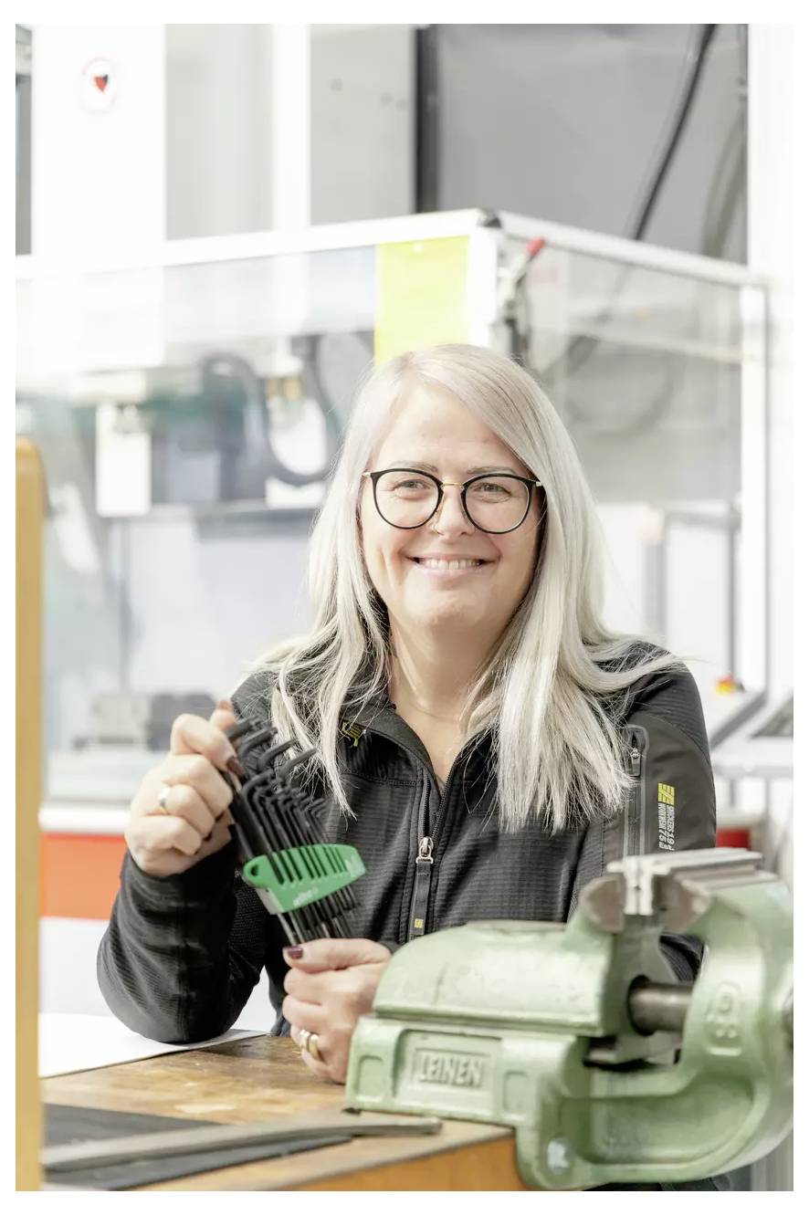 Osoba z długimi włosami i okularami uśmiecha się, trzymając narzędzie. Znajduje się w warsztacie, a w tle widać sprzęt.