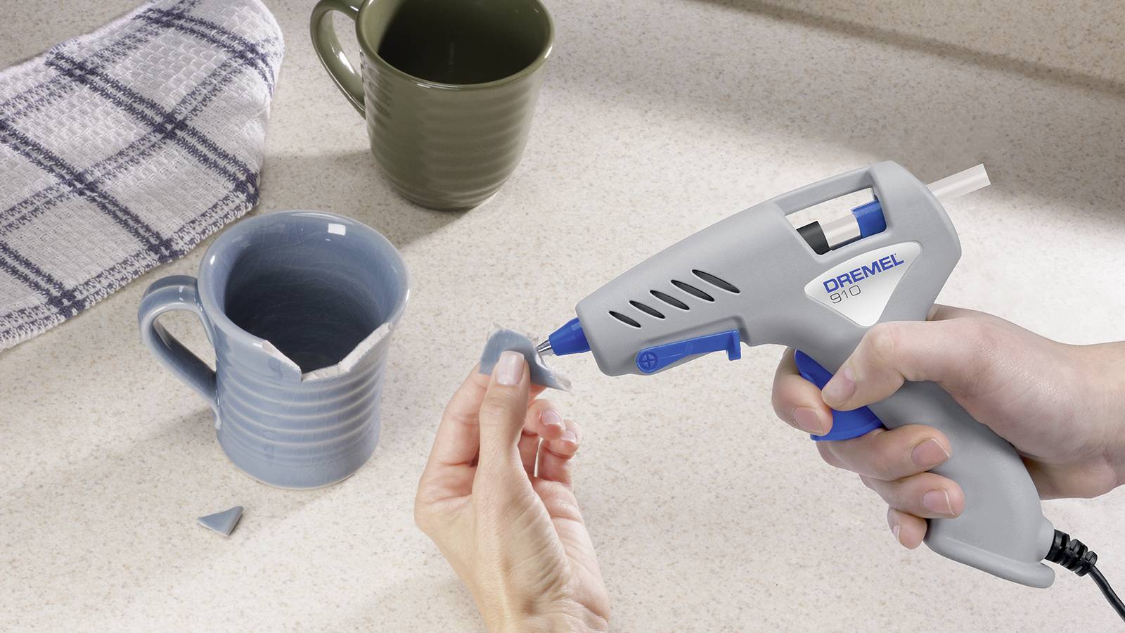 Osoba naprawia złamaną filiżankę za pomocą pistoletu do klejenia na gorąco. W tle widać dwie kolejne filiżanki i ręcznik.