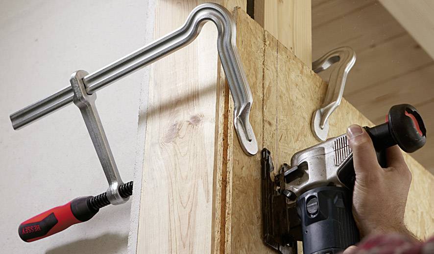 Osoba używa frezarki górnowrzecionowej, aby wykonać wycięcia w desce drewnianej zamocowanej zaciskami śrubowymi do ramy.