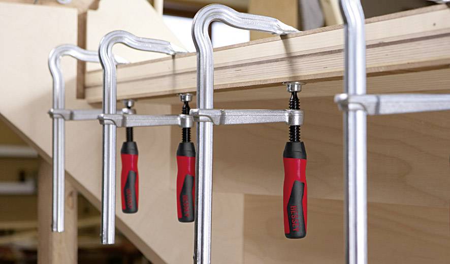 Kilka ścisków śrubowych mocuje ze sobą dwie deski drewniane. Ściski są metalowe z czerwonymi uchwytami i pewnie zabezpieczają deski.