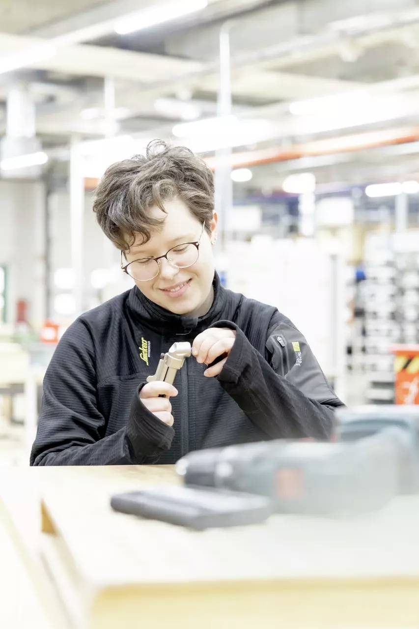 Osoba w okularach używa narzędzia w warsztacie, skupiając się na wykonywanej pracy. W tle widoczne są urządzenia przemysłowe i półki.