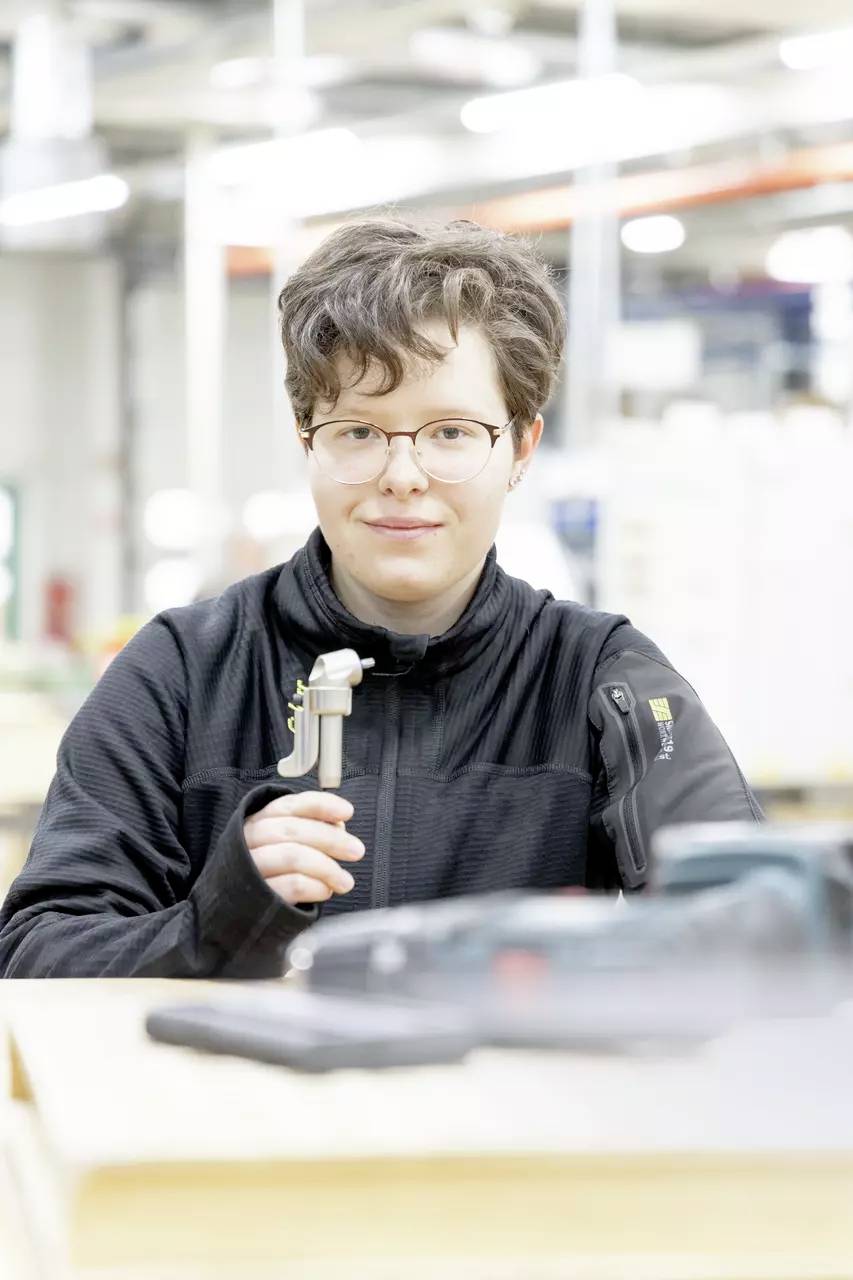 Osoba trzymająca suwmiarkę w warsztacie, w okularach i czarnej kurtce, z rozmytymi narzędziami na pierwszym planie stołu.