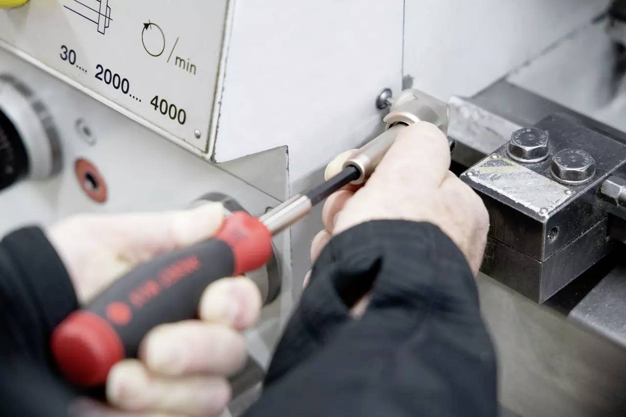 Ręce używające wkrętaka do regulacji części maszyny, obok panelu sterowania przedstawiającego ustawienia prędkości od 30 do 2000 jednostek/min.