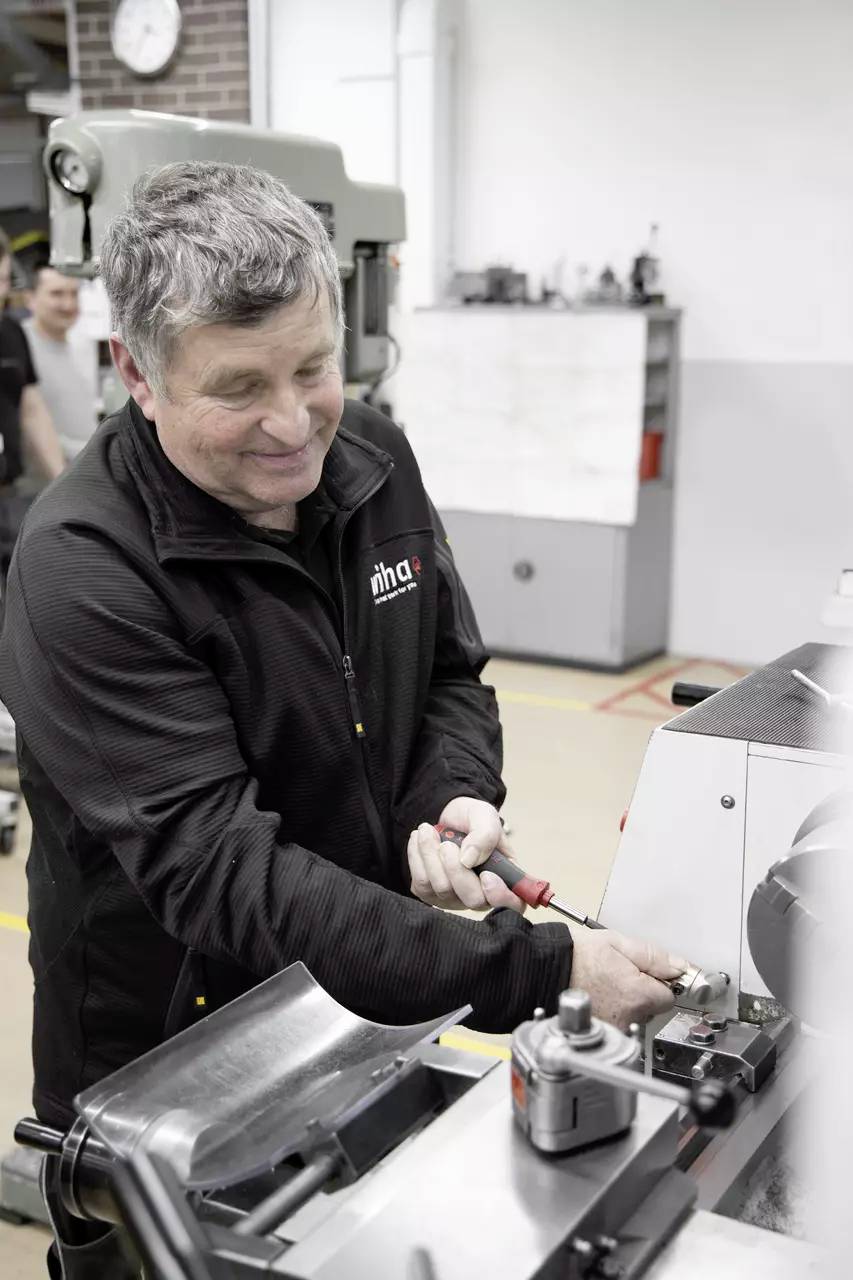 Osoba w warsztacie obsługuje maszynę z skoncentrowanym wyrazem twarzy, ubrana w czarną kurtkę. W tle widoczni są inni pracownicy oraz dodatkowy sprzęt.