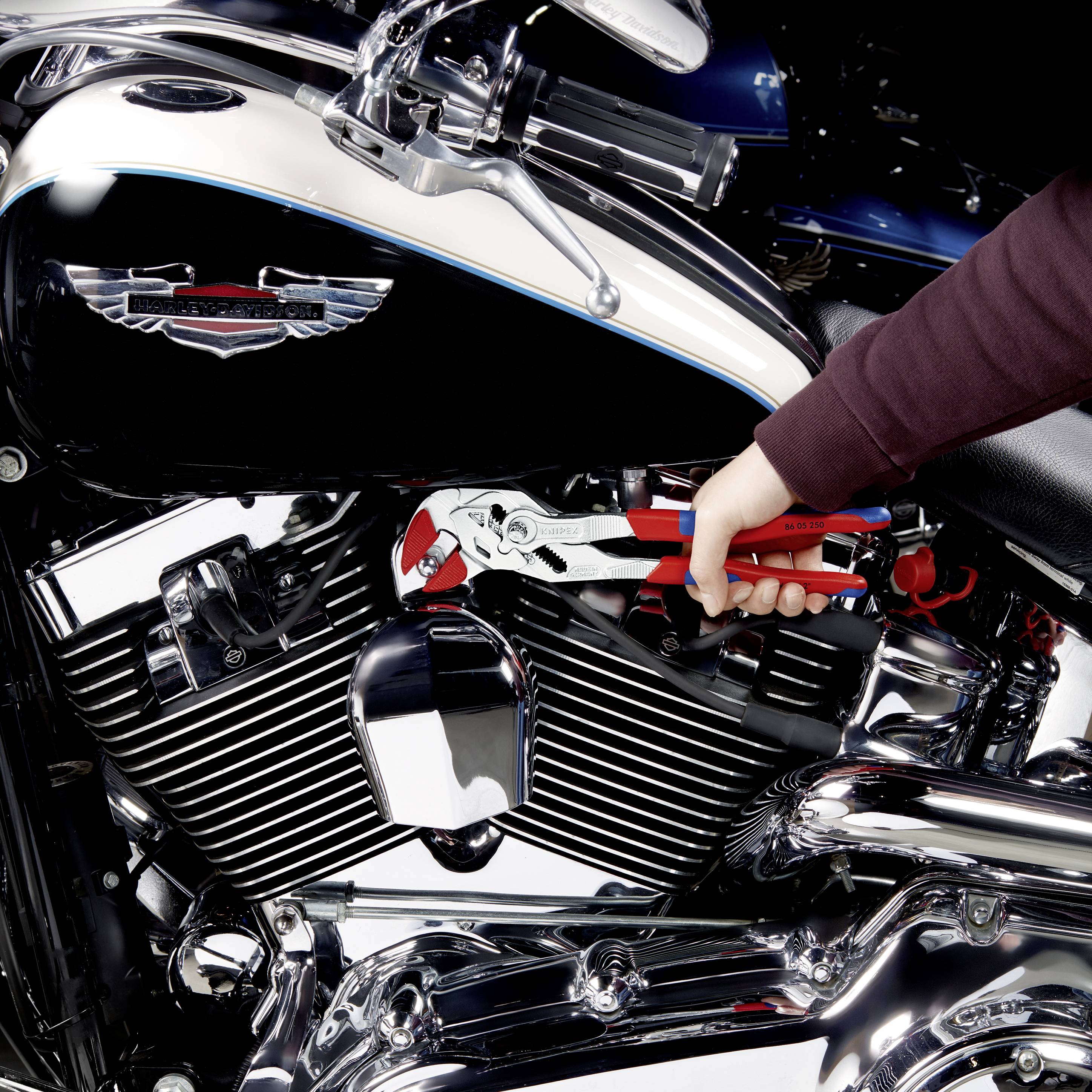 Osoba naprawia motocykl za pomocą szczypiec. Widoczny jest chromowany silnik motocykla i czarny zbiornik paliwa.