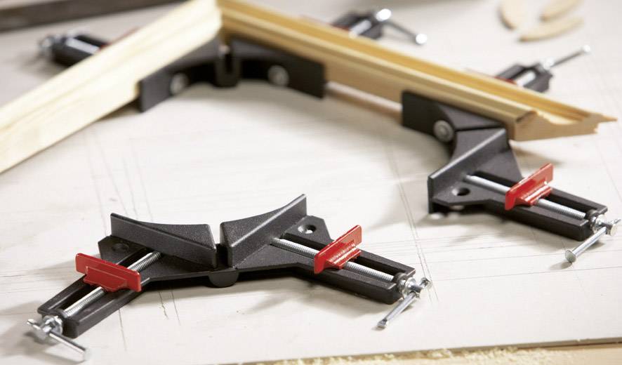 Dwa kawałki drewna są łączone w ramę za pomocą kątowych ścisków stolarskich. Ściski mają czarne uchwyty i czerwone rękojeści.
