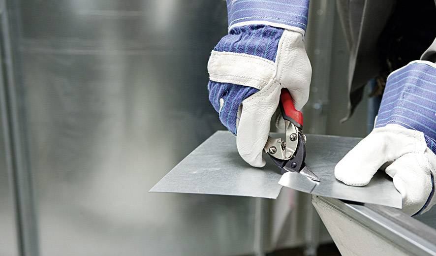 Osoba przecina kawałek blachy metalowymi nożycami. Osoba ma na rękach niebieskie rękawice ochronne.