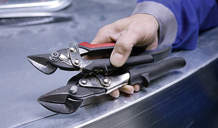 Osoba trzyma dwie nożyce do blachy na metalowej powierzchni. Narzędzia mają ergonomiczne uchwyty w kolorach czarnym i czerwonym.