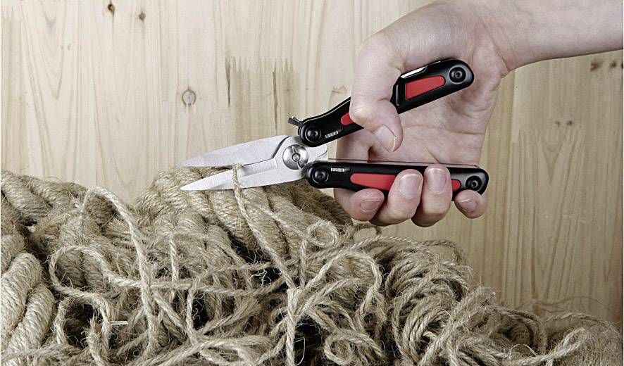 Dłoń trzyma multitool z nożyczkami, który przycina grube sznury na drewnianym tle.