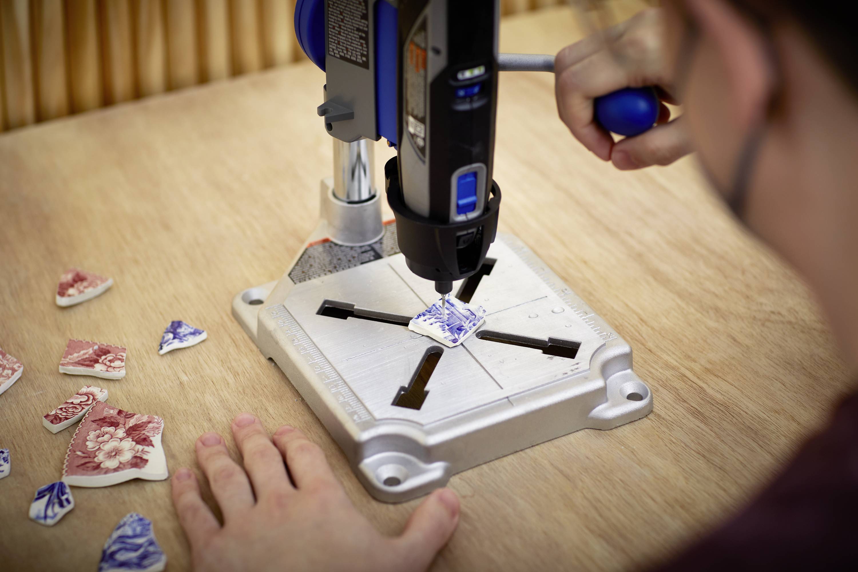 Osoba używa narzędzia Dremel do obróbki ceramicznych odłamków na stole warsztatowym. Kolejne odłamki są rozłożone obok.