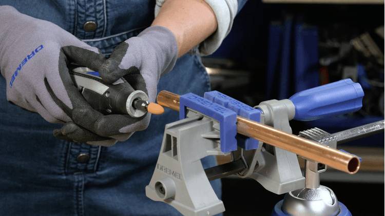 Osoba używa narzędzia obrotowego do polerowania miedzianej rury, która jest zamocowana w imadle na stole warsztatowym. Ma na sobie rękawice i dżinsową koszulę.