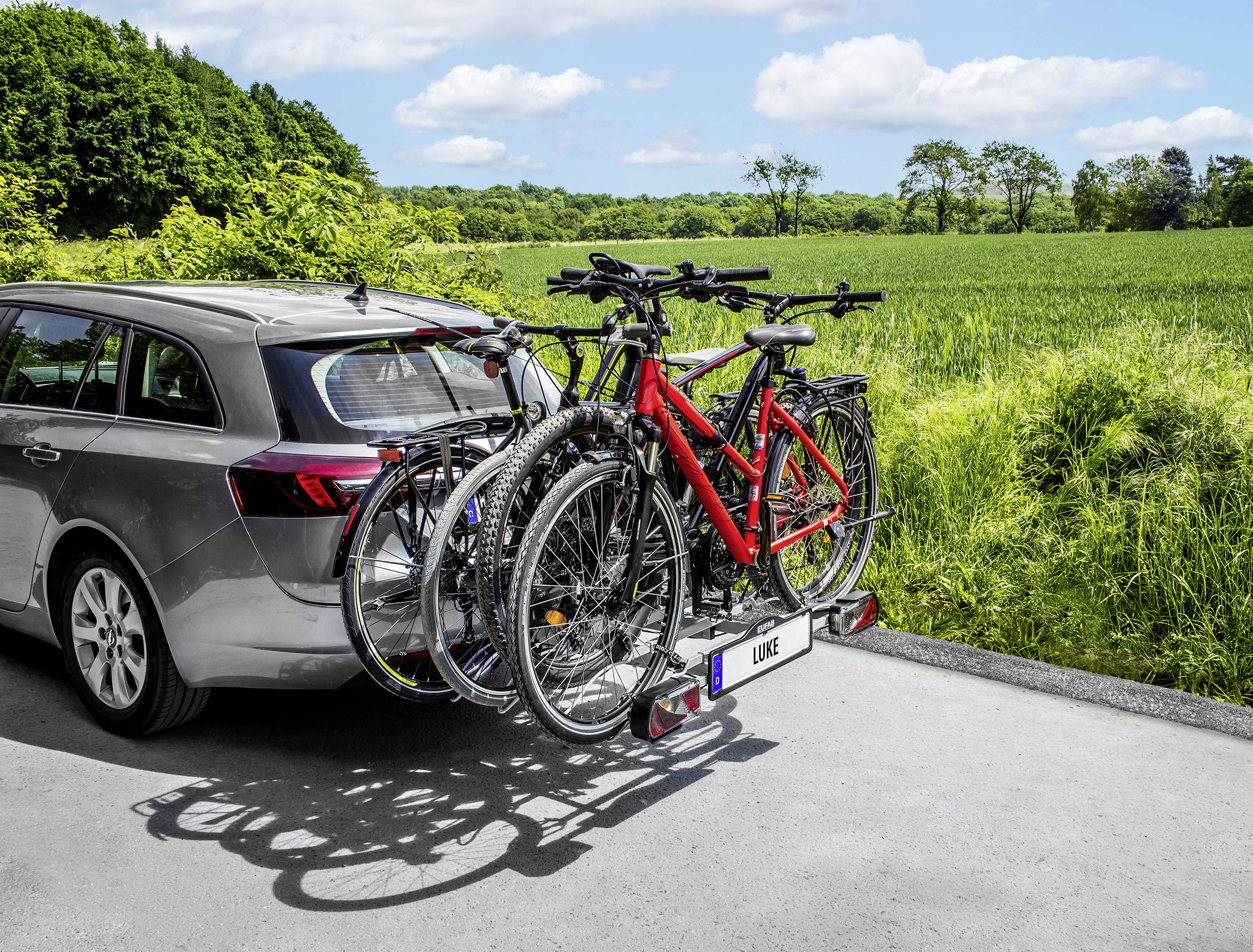 Srebrny kombi z bagażnikiem rowerowym przewozi cztery rowery drogą krajową; w tle widać pola i drzewa.