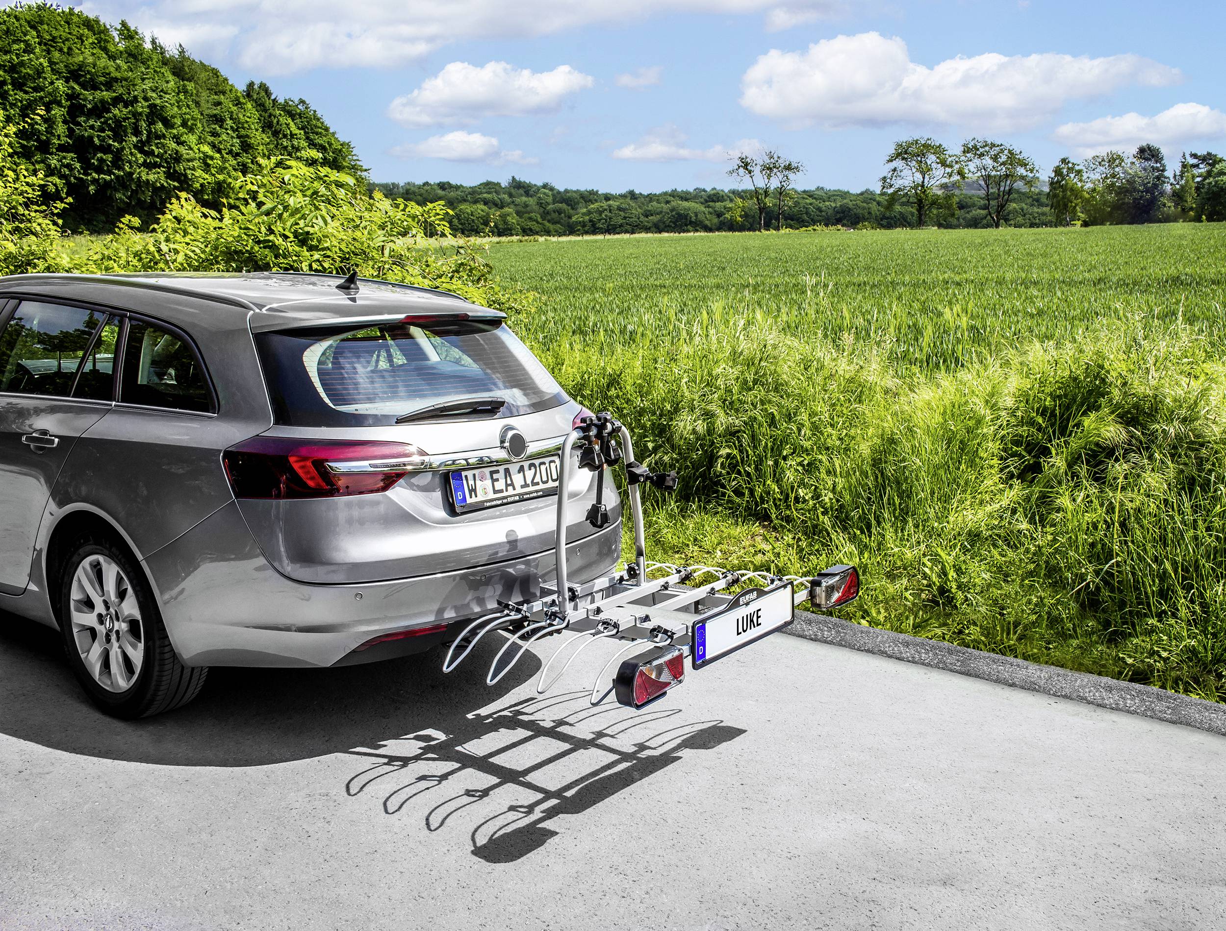 Srebrny kombi zaparkowany przy krawędzi drogi z bagażnikiem rowerowym z tyłu. W tle zielone pola i drzewa pod niebieskim niebem.