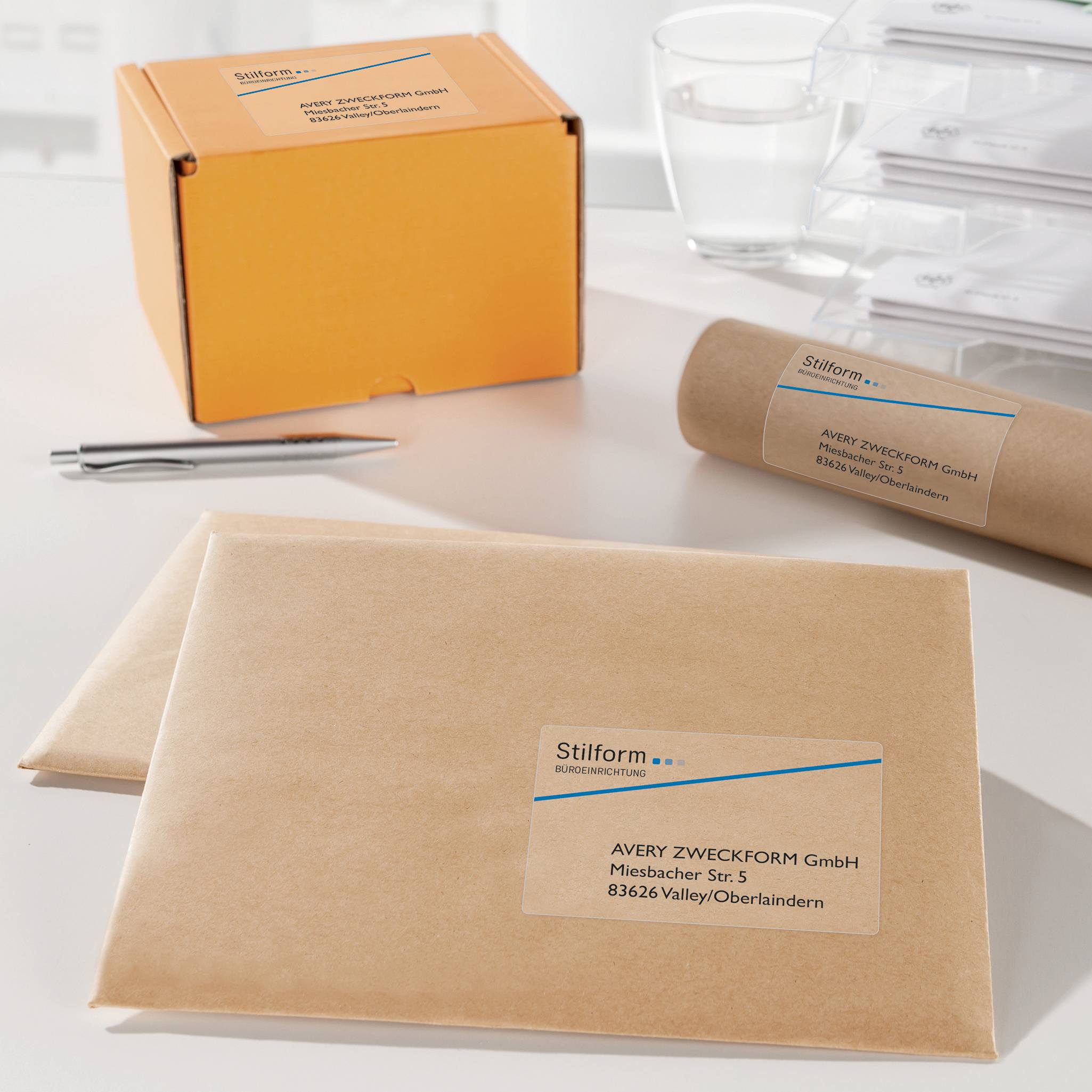 Paczki z etykietami adresowymi i logo firmy 'Stilform' na białym stole, obok długopis i szklanka wody.