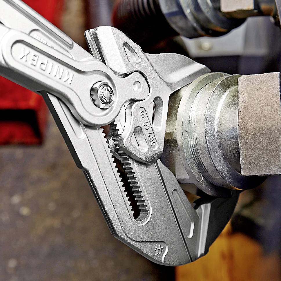 Szara szczypce z prążkowaną krawędzią uchwytu leżą otwarte na metalowym elemencie. Widoczna marka szczypiec. Miejsce pracy ma przemysłowy charakter.