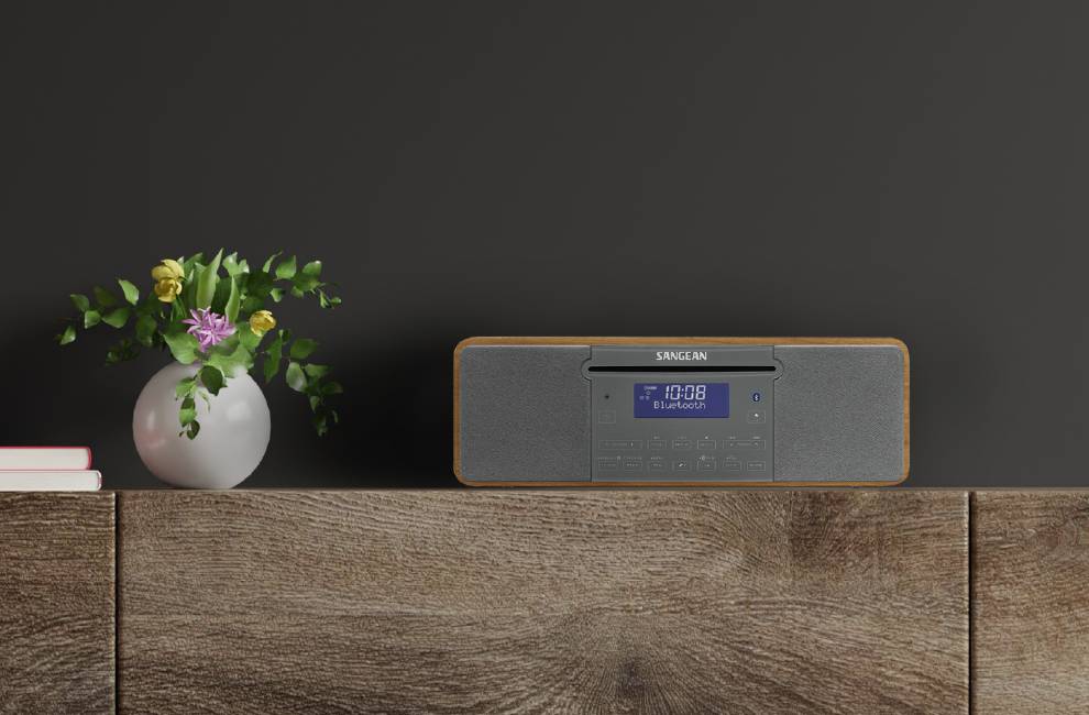 Drewniany stół z radiem i wyświetlaczem cyfrowym pokazującym '10:08'. Obok stoi wazon z kolorowymi kwiatami.