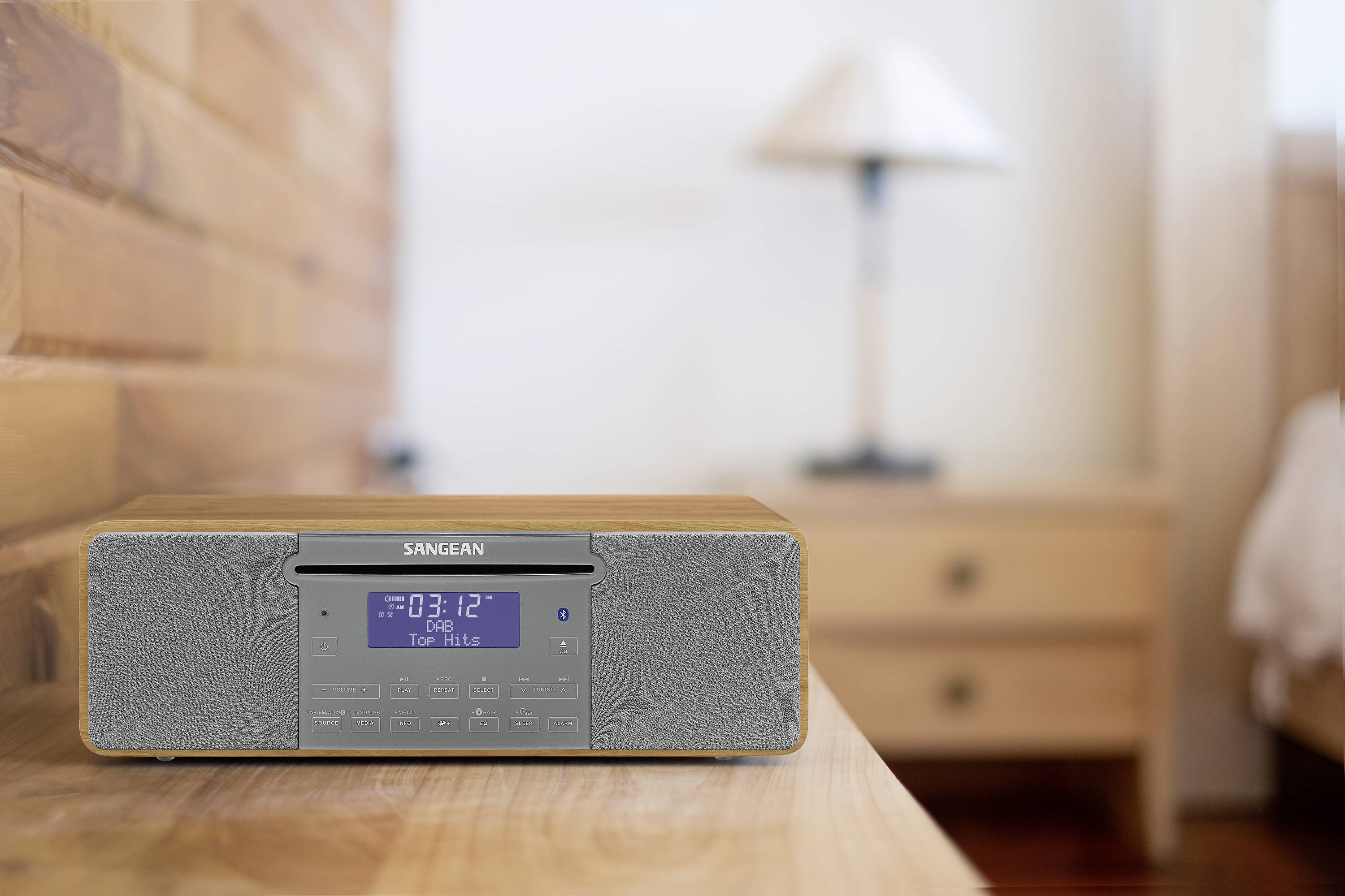 Radio na drewnianym stole na pierwszym planie, z wyświetlaczem cyfrowym pokazującym godzinę '08:12'. W tle widać sypialnię.