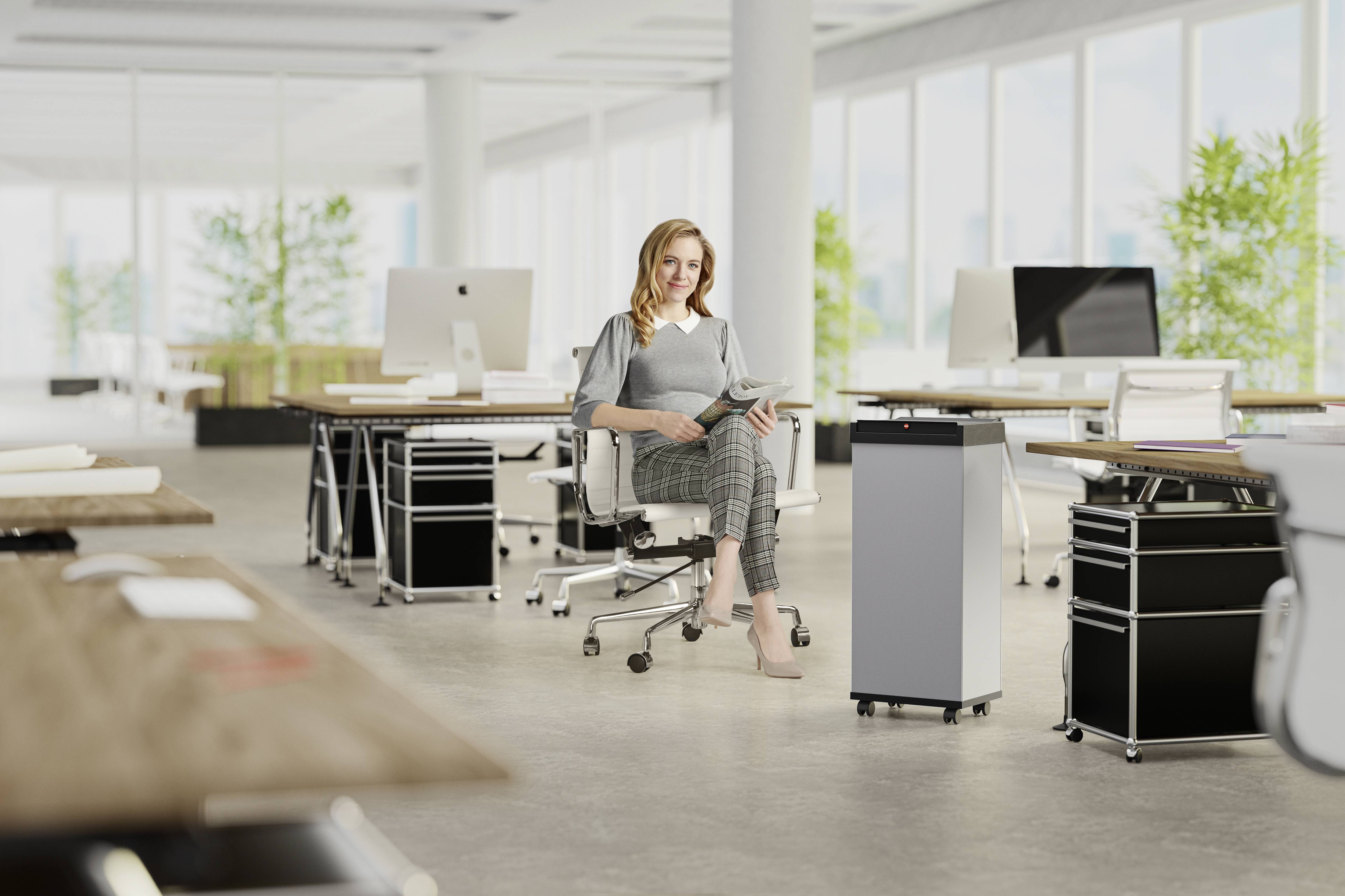 Kobieta siedzi z tabletem w nowoczesnym biurze z wieloma stanowiskami pracy. Duże okna w tle wpuszczają dużo światła.