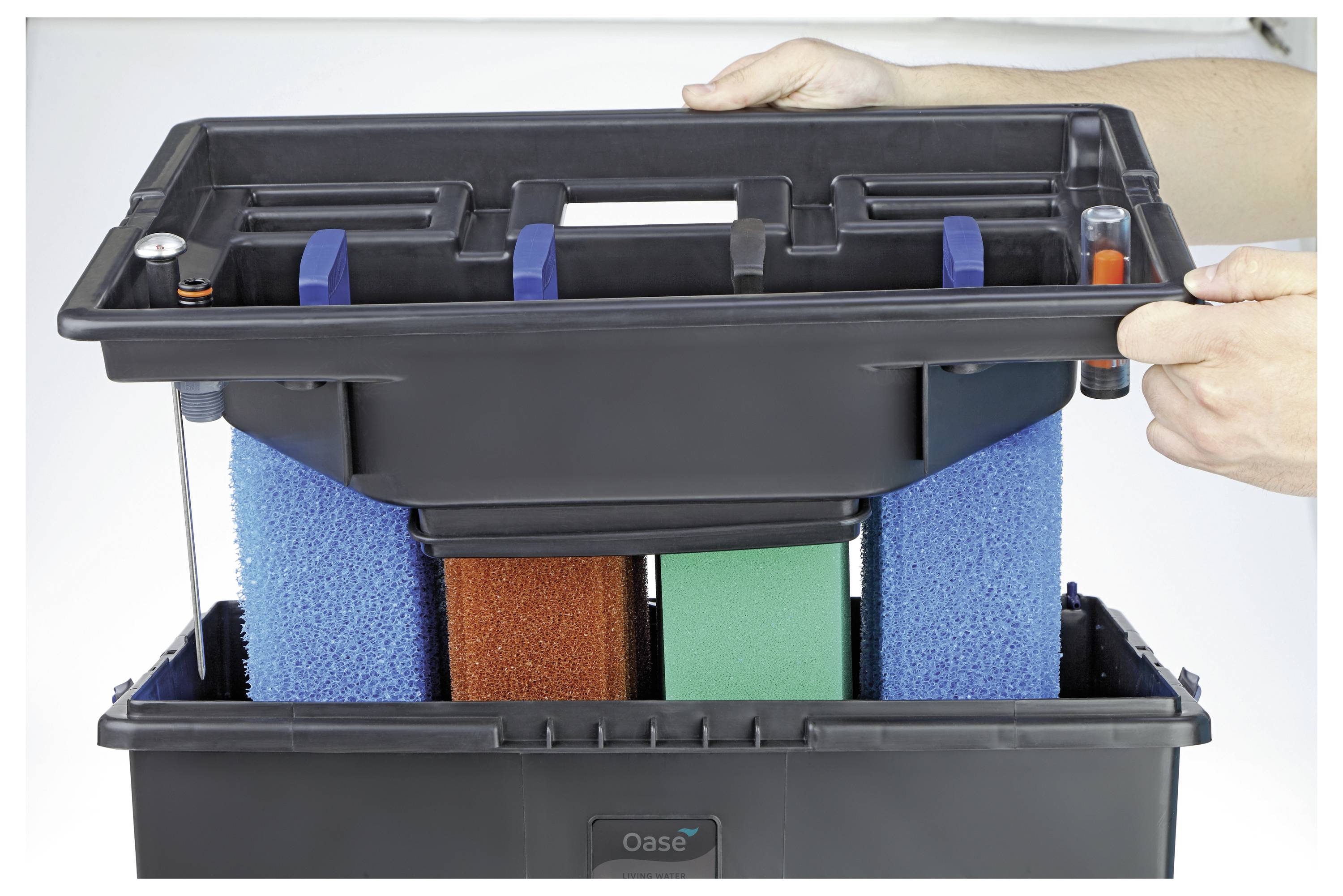 Ręce podnoszące górną sekcję czarnej jednostki filtra akwariowego, odkrywające wielobarwne gąbki filtracyjne poniżej podczas czynności konserwacyjnych.