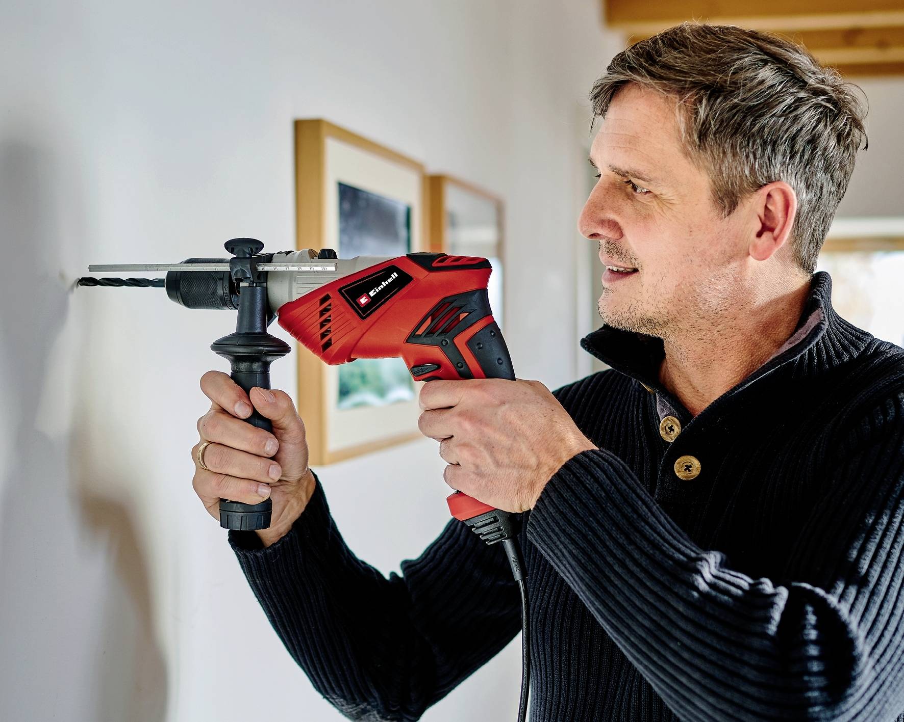 Mężczyzna wiertarką w kolorze czerwonym wywiera otwór w białej ścianie. W tle widoczne są oprawione obrazy.