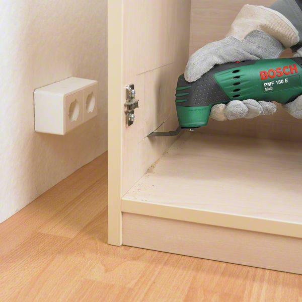 Osoba używa wielofunkcyjnego narzędzia Bosch PMF 180 E do obróbki drewnianej półki. Gniazdo elektryczne jest widoczne na ścianie.