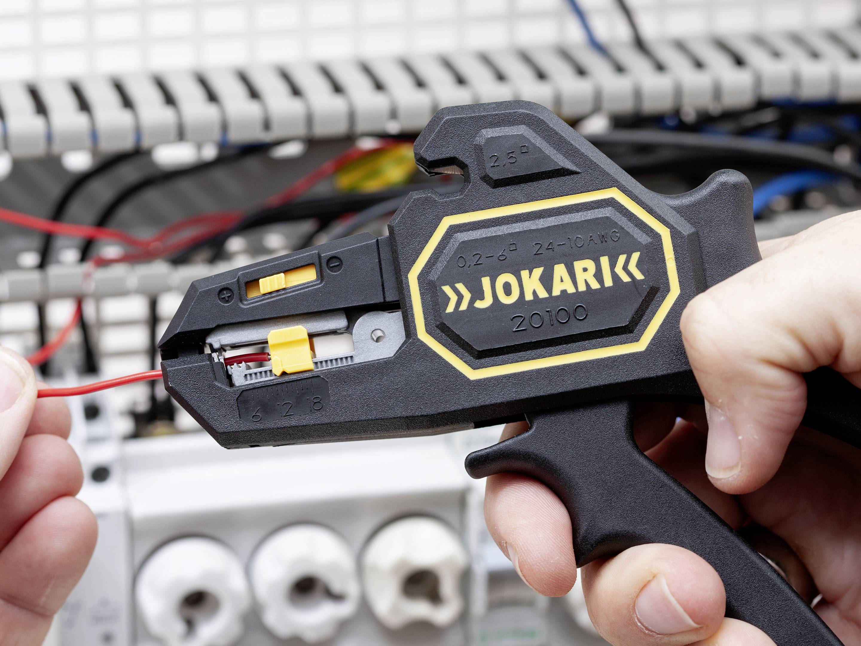 Osoba używa szczypiec Jokari do zdejmowania izolacji z czerwonego przewodu w szafce elektrycznej.