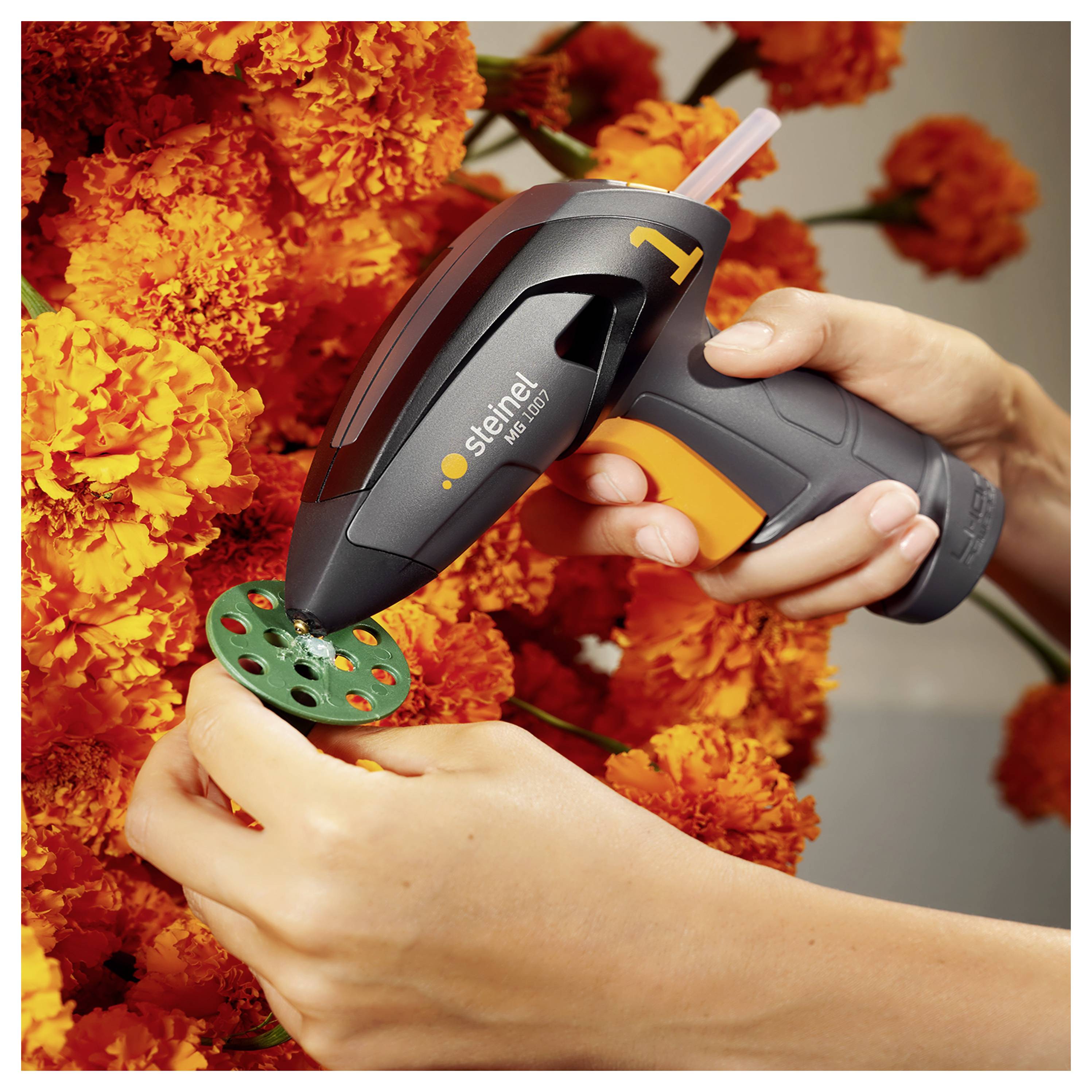 Ręce używające pistoletu do kleju Steinel do przytwierdzenia zielonego elementu rzemieślniczego wśród pomarańczowych kwiatów.