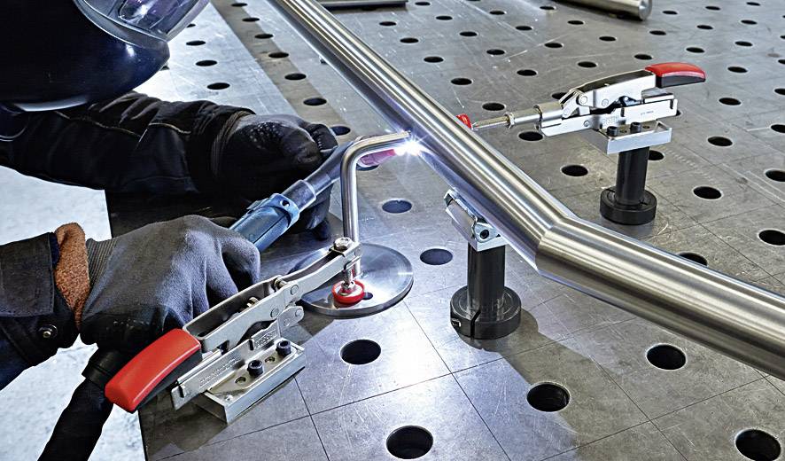 Osoba spawająca metalową rurę na stole roboczym w czarnej spawalniczej rękawicy i kasku. Metalowe zaciski przytrzymują rurę.