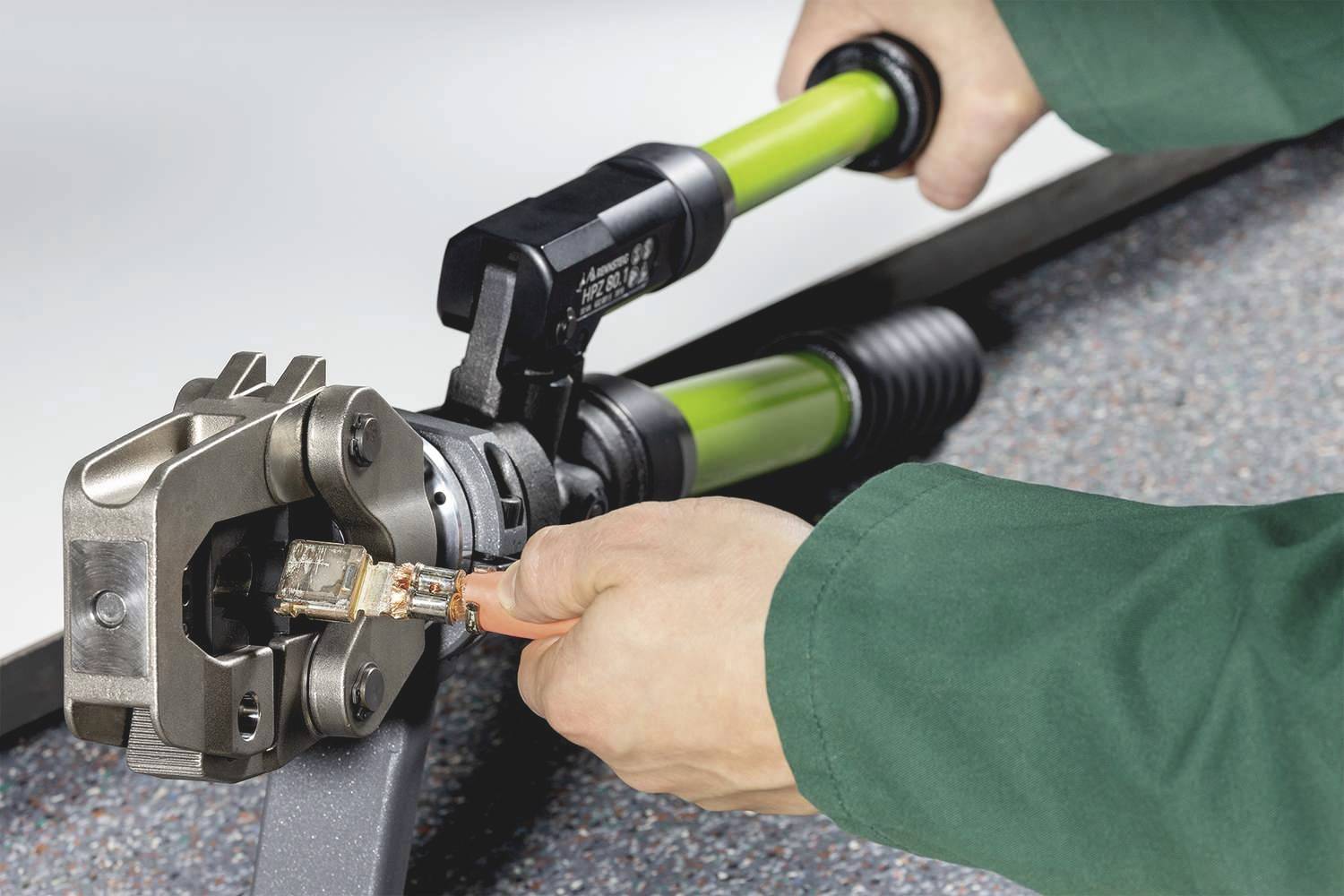 Osoba w zielonym płaszczu używa szczypiec hydraulicznych do zaciskania wtyku RJ45 na kablu sieciowym.