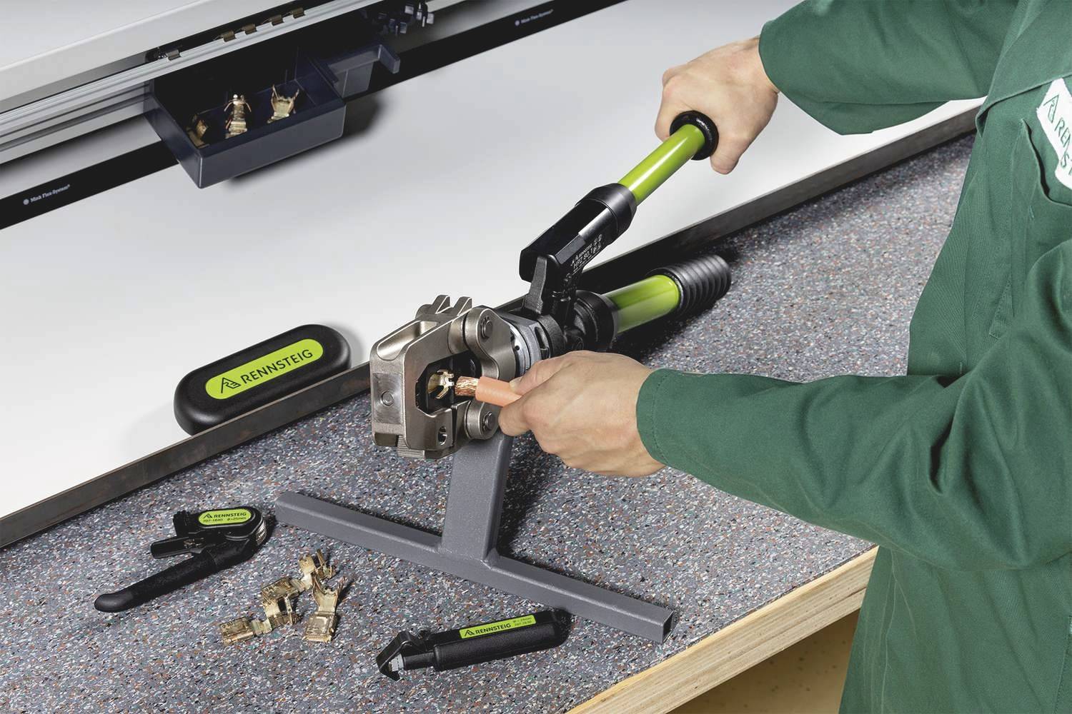 Osoba ubrana w zieloną odzież roboczą używa narzędzia z długą rączką, aby zamocować metalowy element na stole warsztatowym.