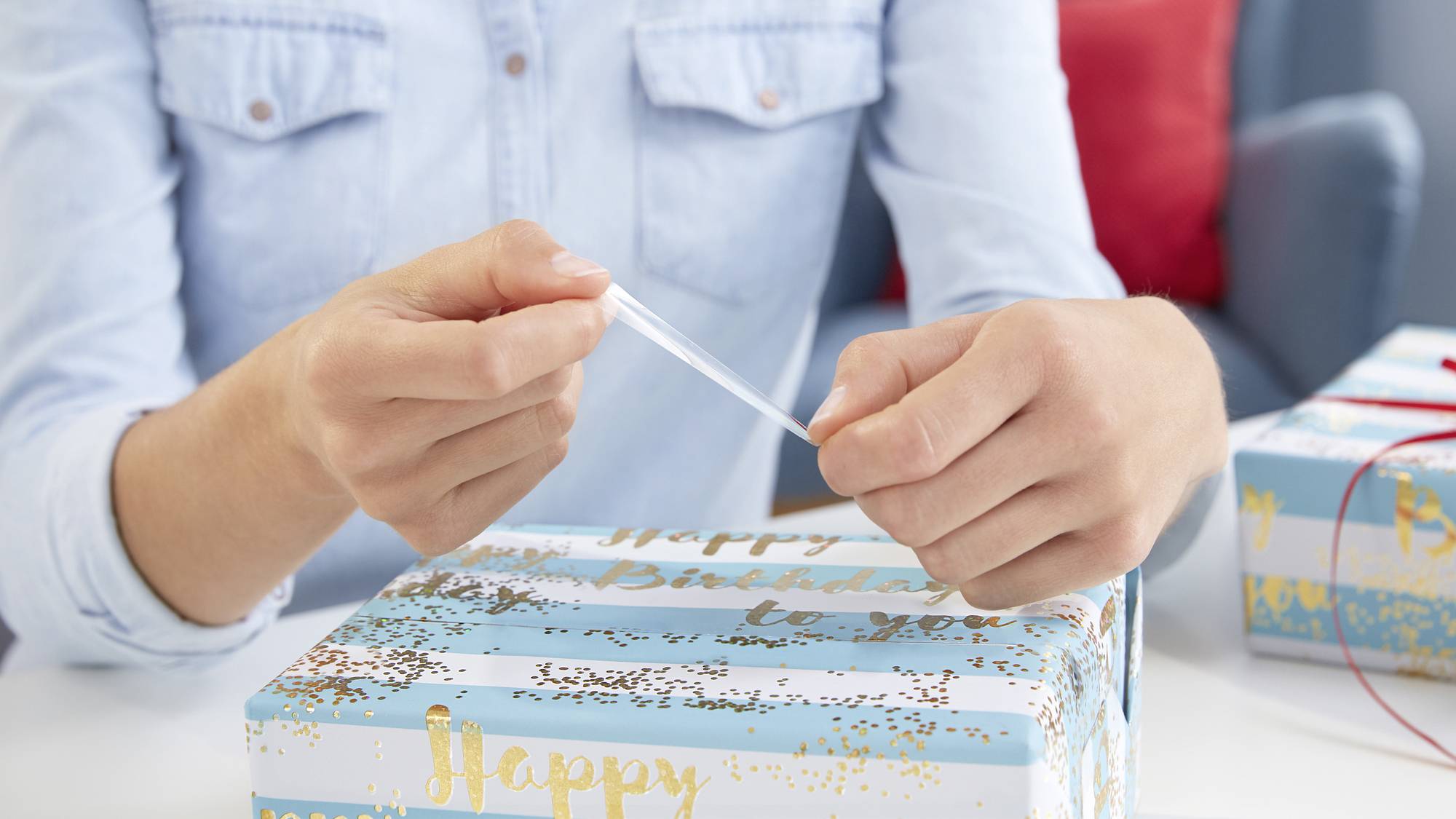 Osoba pakuje prezent w papier w kolorach niebieskim, białym i złotym, na którym widnieje napis „Happy Birthday to you".