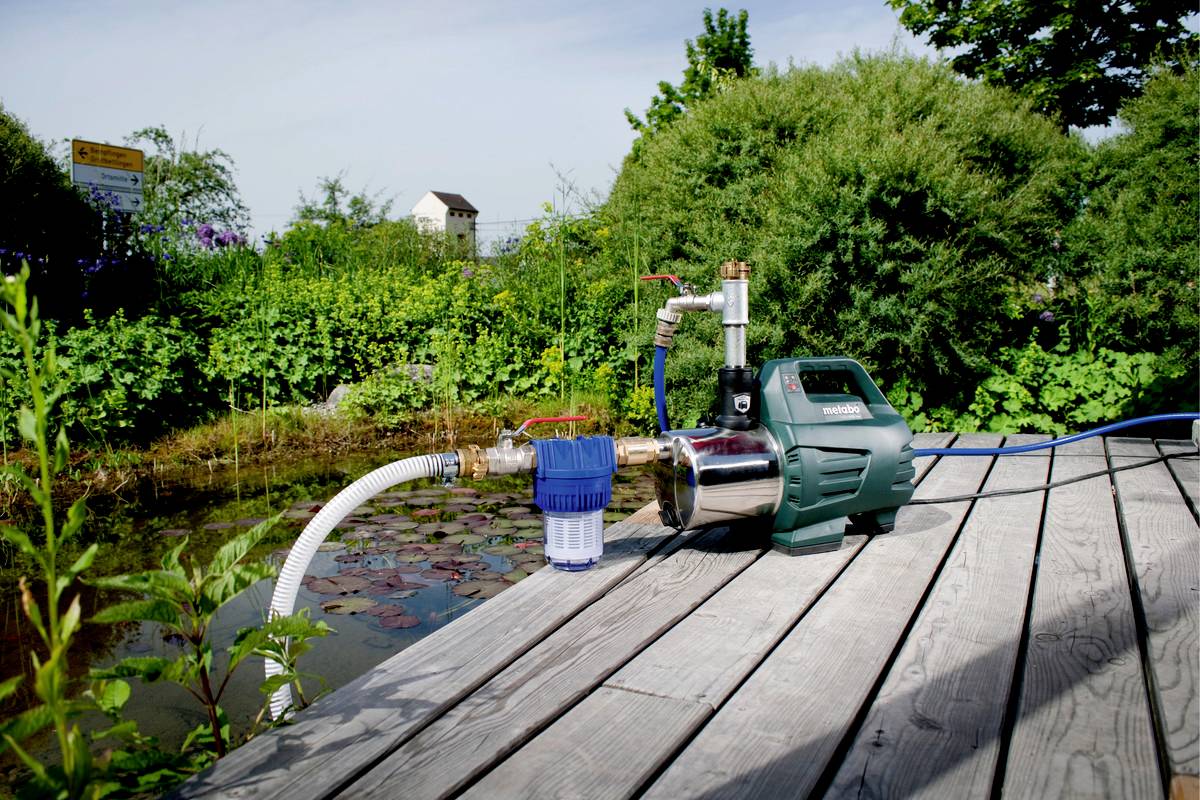 Pompa wodna na drewnianym tarasie obok stawu, otoczona zielonymi roślinami. Pompa posiada niebieski system filtracyjny.