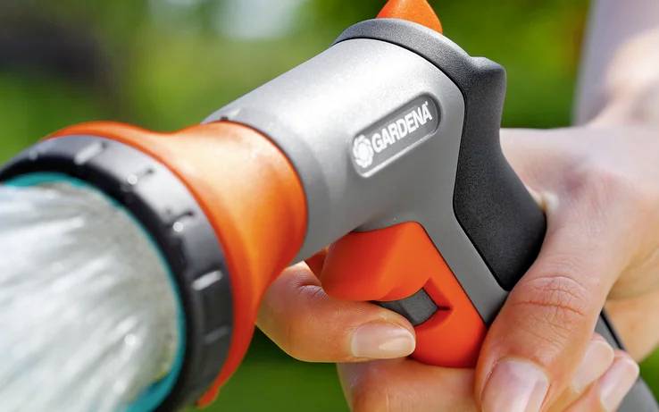 Osoba reguluje dyszę węża ogrodowego, z której tryska woda. Dysza jest szara z pomarańczowymi akcentami, trzymana w dłoni na zewnątrz.