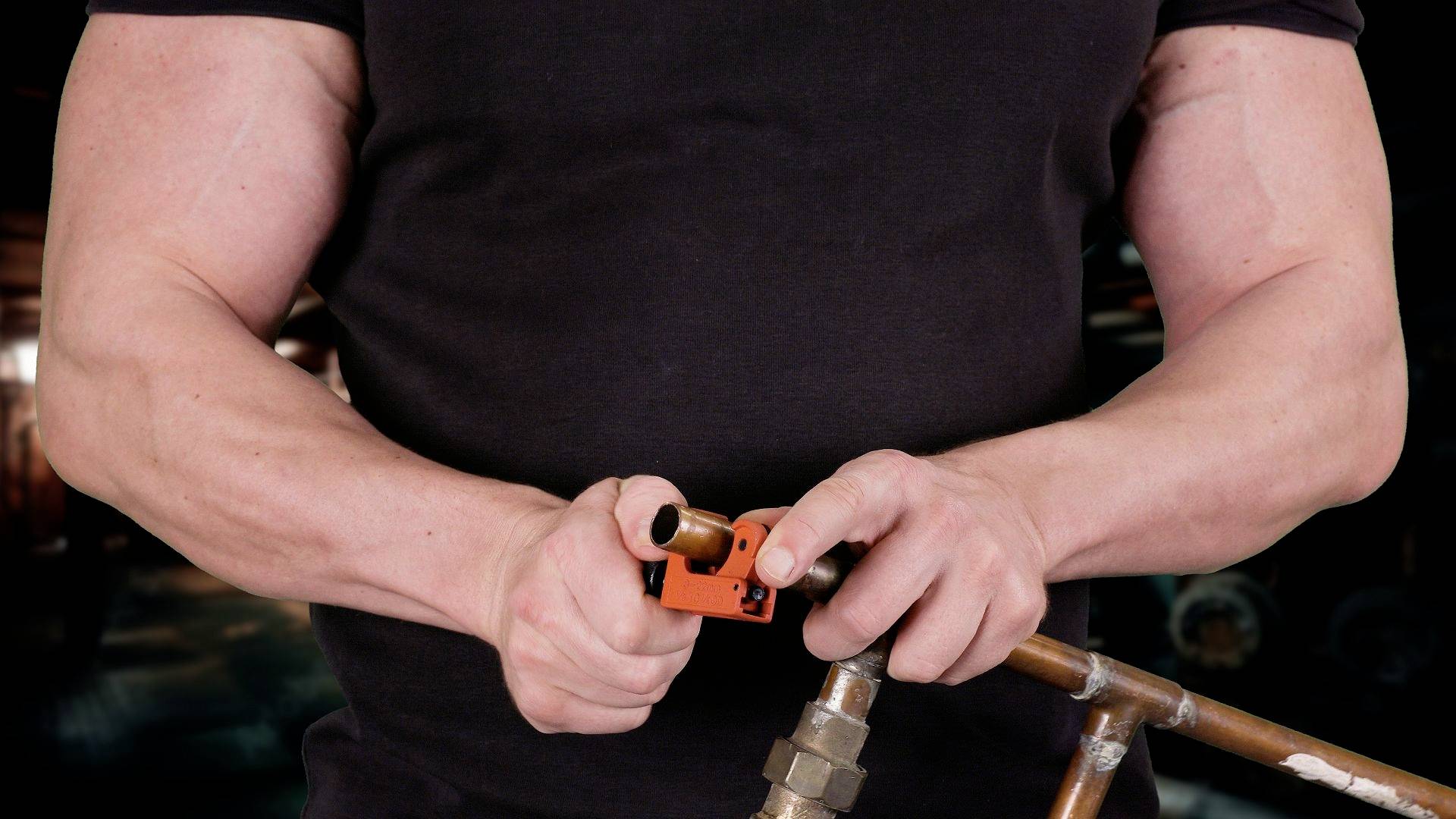 Umięśniona osoba w czarnym t-shircie używa narzędzia do cięcia miedzianej rury w warsztacie.