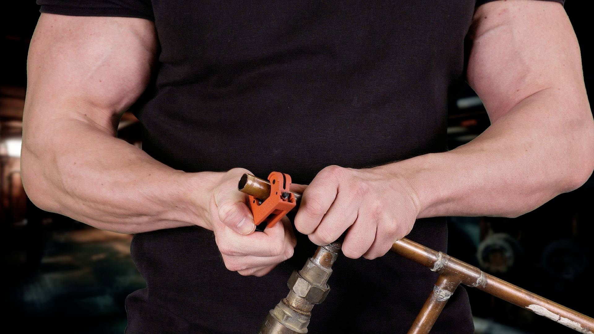 Osoba przecina rurę miedzianą za pomocą obcinaka do rur. Otoczenie wskazuje na warsztat lub plac budowy.