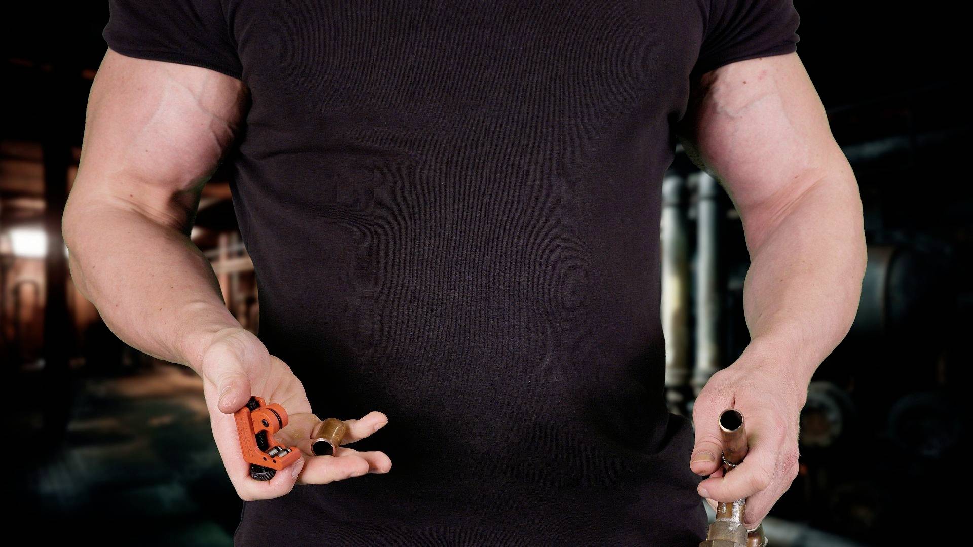 Osoba w czarnym t-shircie trzyma narzędzia w obu dłoniach. Tło przypomina warsztat lub środowisko przemysłowe.