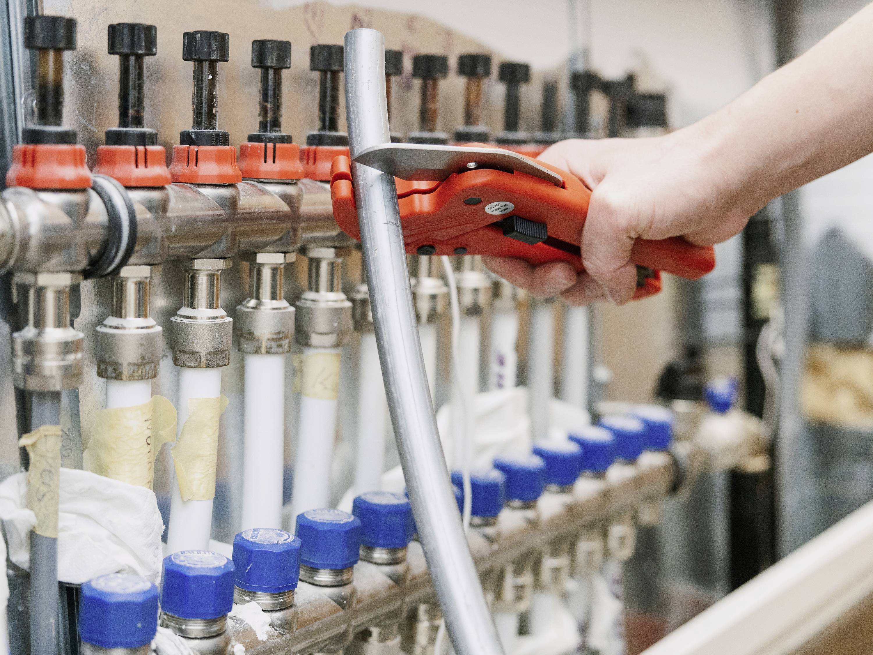 Osoba używa czerwonego obcinaka do rur, aby przeciąć metalową rurę przy zestawie zaworów i przewodów w pomieszczeniu technicznym.