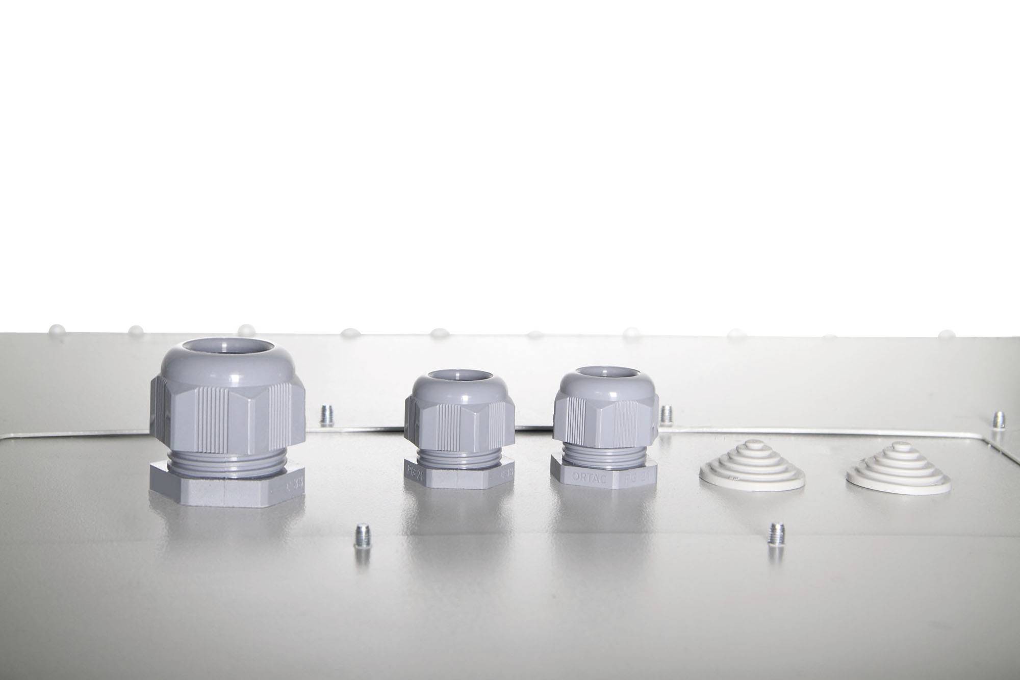 Trzy szare dławnice kablowe o różnych rozmiarach na metalowej powierzchni, obok których znajdują się dwa białe obiekty o kształcie stożkowym.