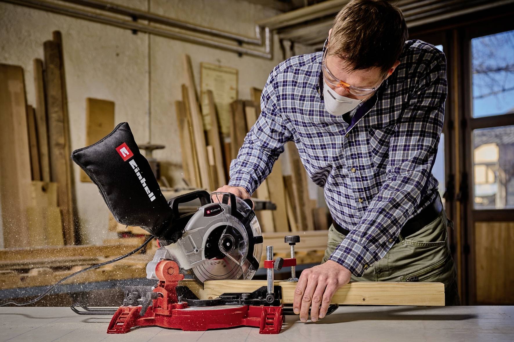 Mężczyzna bezpiecznie pracuje piłą tarczową, nosząc okulary ochronne i maskę przeciwpyłową. W tle widać narzędzia do obróbki drewna.