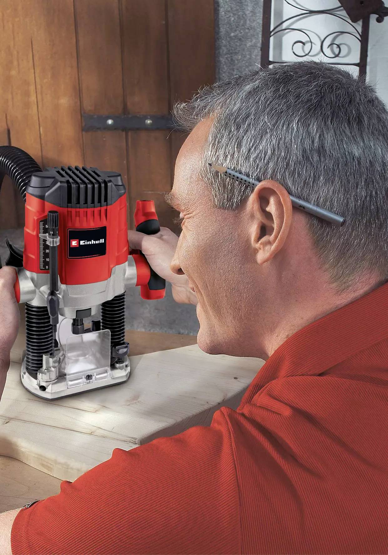 Osoba używa czerwonej frezarki na desce drewnianej, mając ołówek za uchem.