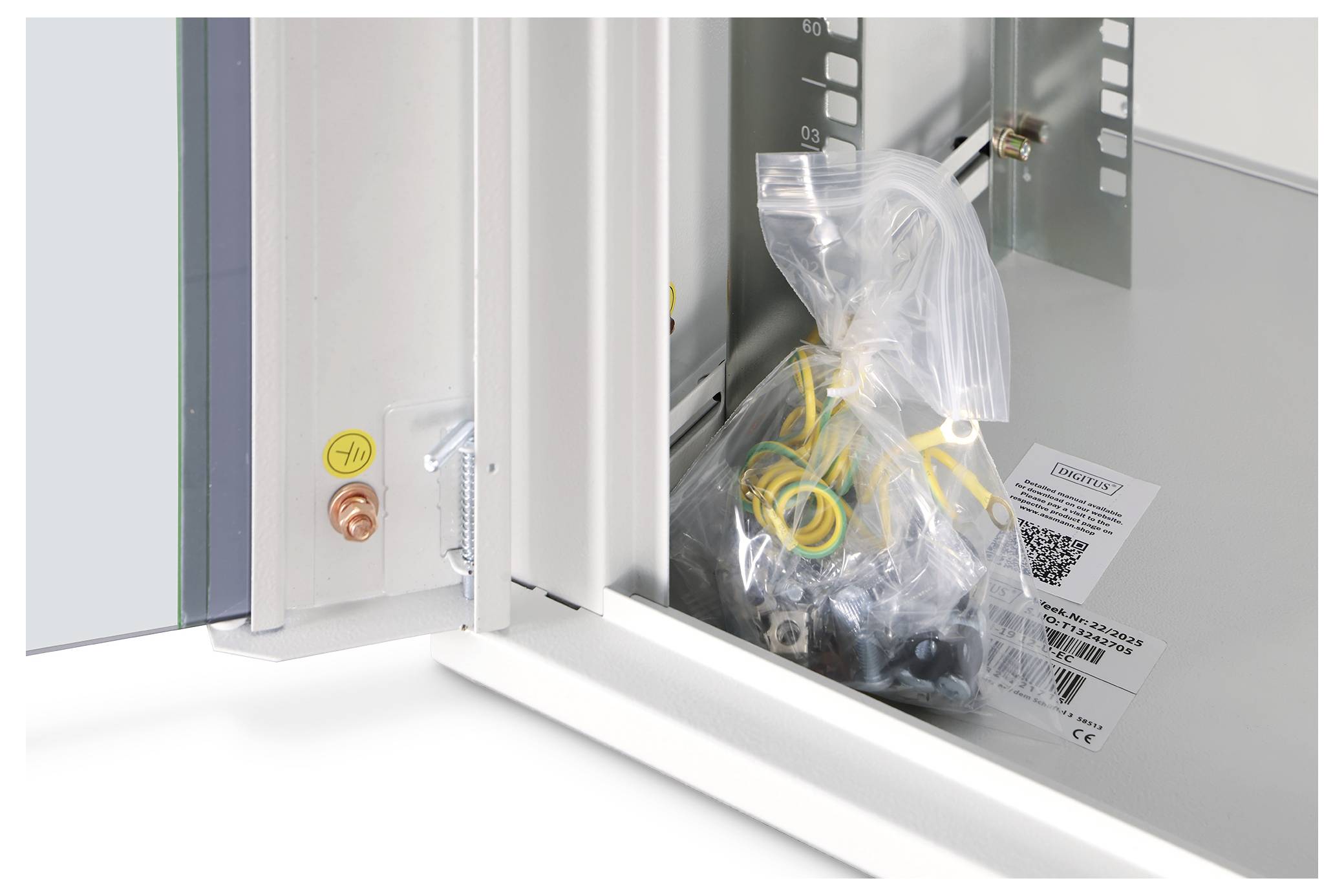 Otwarta szafka prezentuje plastikową torbę zawierającą przewody i komponenty. Wydrukowana etykieta z kodem QR znajduje się obok torby.