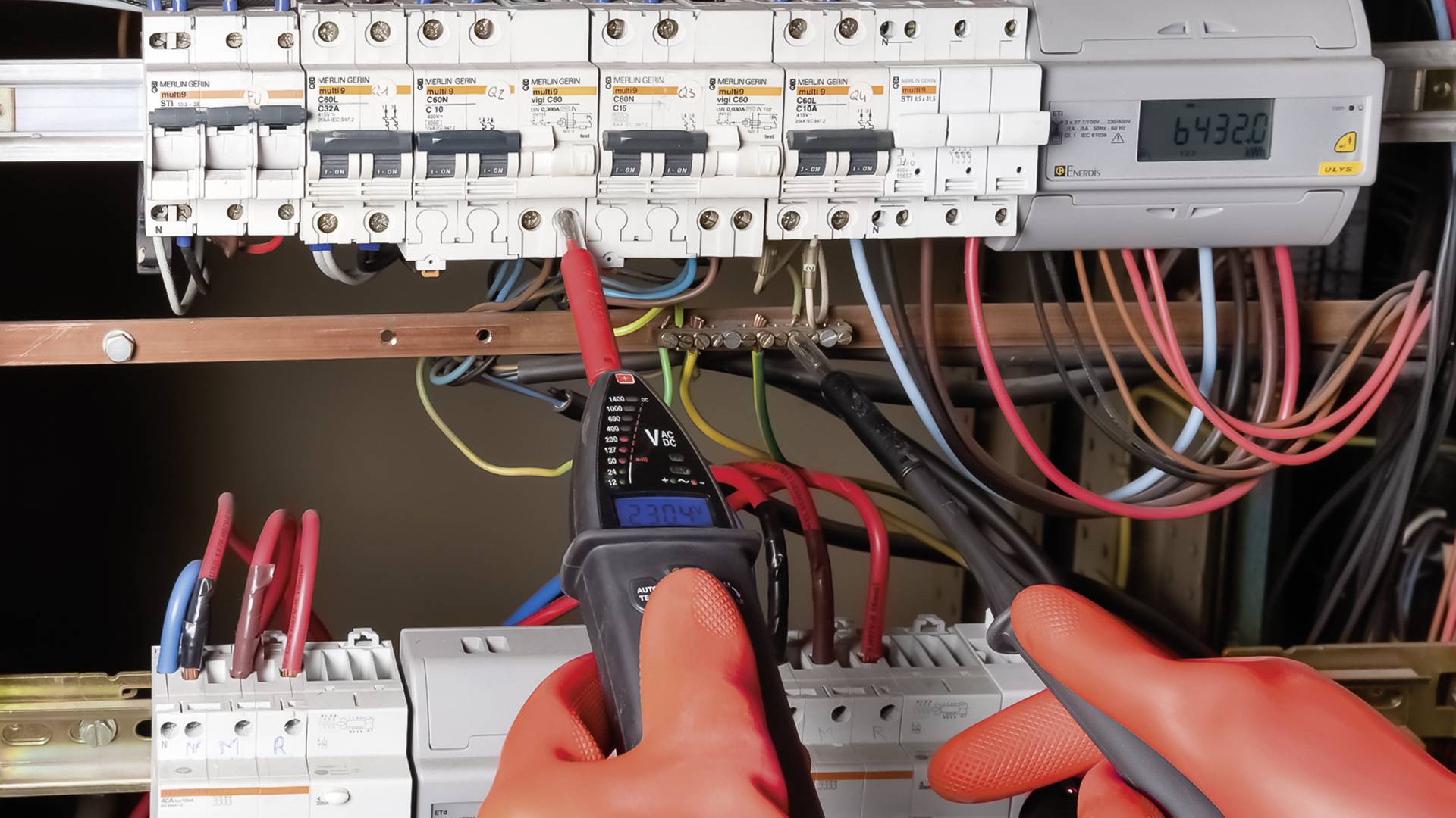 Elektryk sprawdza napięcie za pomocą miernika w skrzynce bezpiecznikowej. Liczne kolorowe przewody są podłączone. Wyświetlacz pokazuje '64320'.